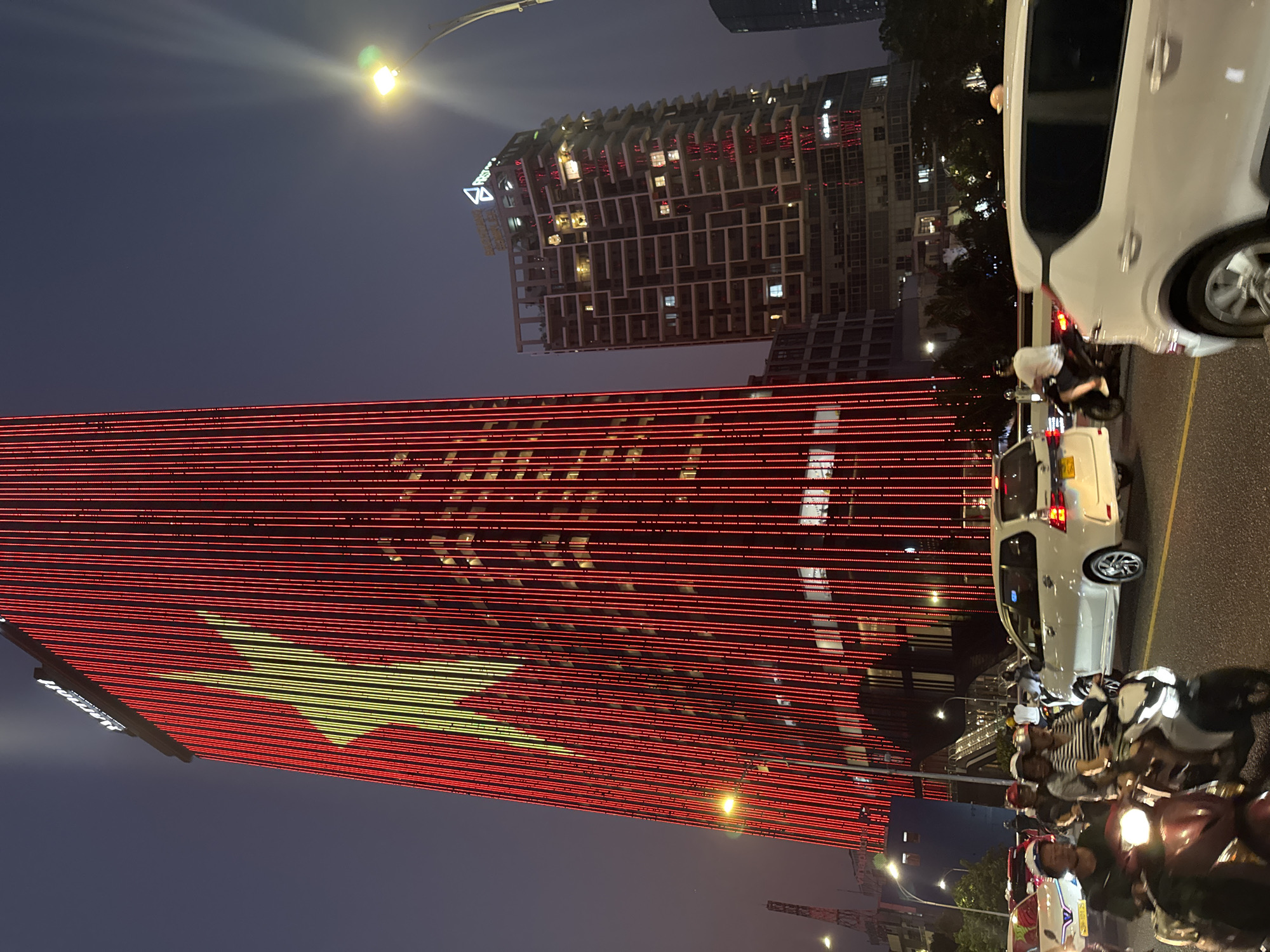 The image shows a night scene in Đà Nẵng, Vietnam. A building is decorated with red horizontal lights arranged to form the Vietnamese flag, with a yellow star. Several other buildings are visible in the background, including one with a distinctive maze-like facade. In the foreground, a street is filled with motorbikes and cars waiting at what is probably a stoplight or an intersection. The road appears to be asphalt. A street light illuminates the scene from above.