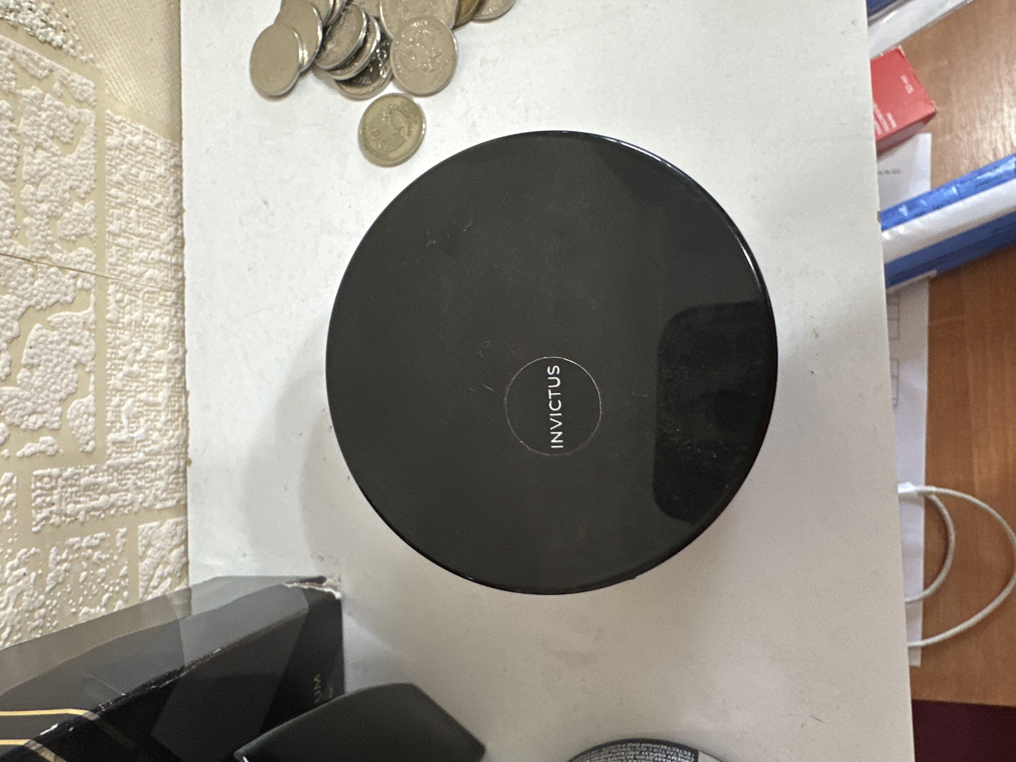 In Rostov-on-Don, Russia, on a textured, light-colored surface that appears to be a desk or countertop, a black circular container with the inscription "INVICTUS" in white lettering is centrally positioned. To the right of the container, a pile of silver-colored coins is visible, some stacked and others lying flat. In the background, a textured, off-white wall with a repeating pattern can be observed. To the left of the container, a portion of a black object, possibly a box or packaging with faint text, is present. The overall scene is an indoor, static arrangement of objects on a flat surface.