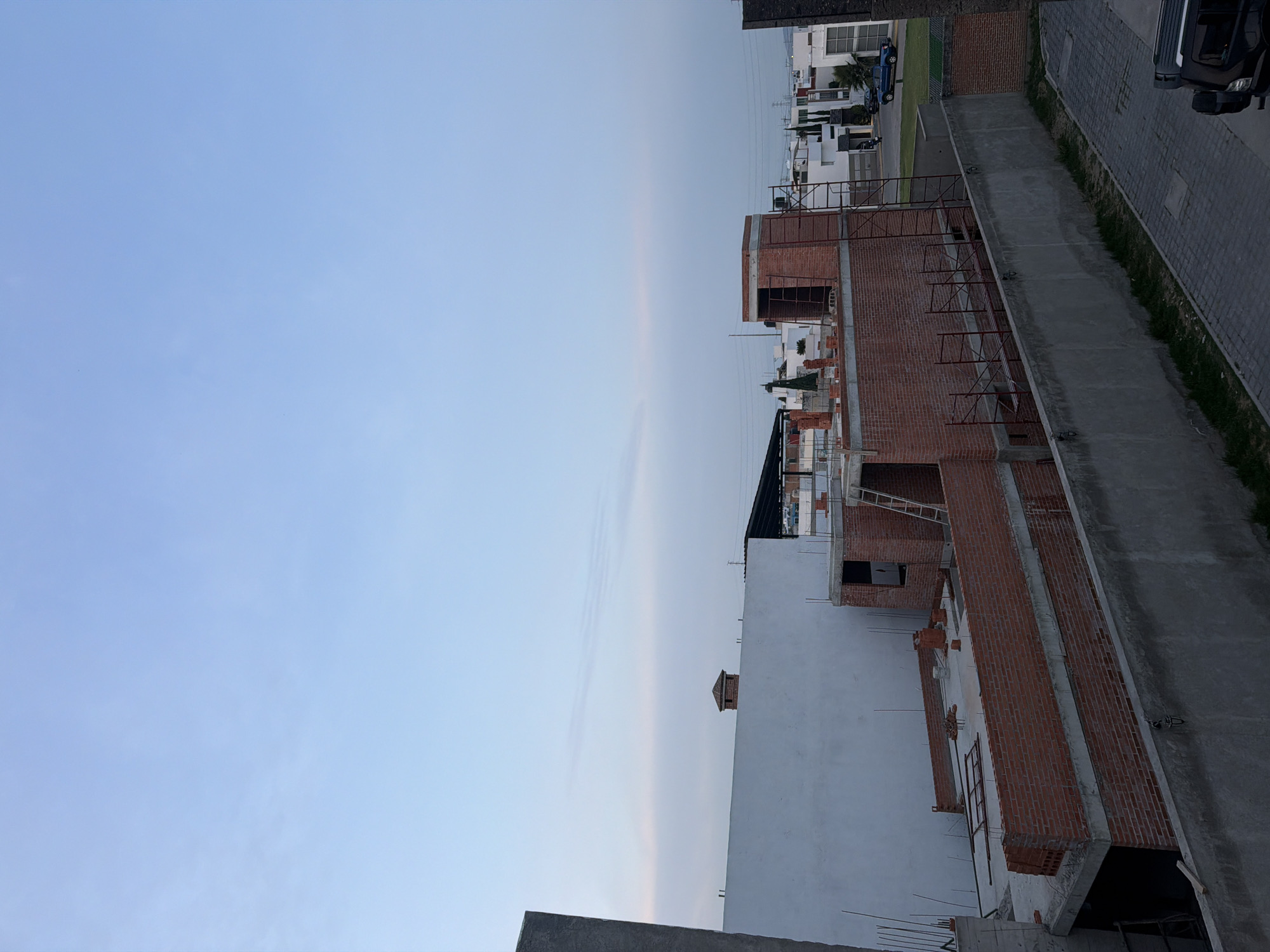 The image captures a panoramic view of an urban or suburban landscape in San Andrés Cholula, Mexico, characterized by a mix of ongoing construction and established residential areas under a soft, light blue sky.
In the foreground, a dark, flat concrete or rooftop structure is partially visible at the bottom. The mid-ground is dominated by several buildings in various stages of construction. These structures prominently feature exposed reddish-brown brick walls and grey concrete elements. Some sections of the buildings have already been painted white. Metal scaffolding or support structures are attached to the unfinished brickwork, and a ladder is propped against one of the taller constructions.
To the right of the construction site, a paved road or street stretches alongside the buildings. A dark-colored SUV or truck is partially visible, parked on or near this road. Further in the background, beyond the immediate construction, a row of completed, modern white residential houses with dark roofs and prominent windows can be seen, interspersed with green landscaping and trees. Faint power lines are also visible against the distant horizon.
The upper half of the image is filled with a vast, clear, pale blue sky, suggesting either early morning or late afternoon/dusk. The lighting is soft and even, with no harsh shadows, implying a calm weather condition. No people are visible, and there is no discernible text within the frame. The overall scene depicts an active area of residential development and expansion.