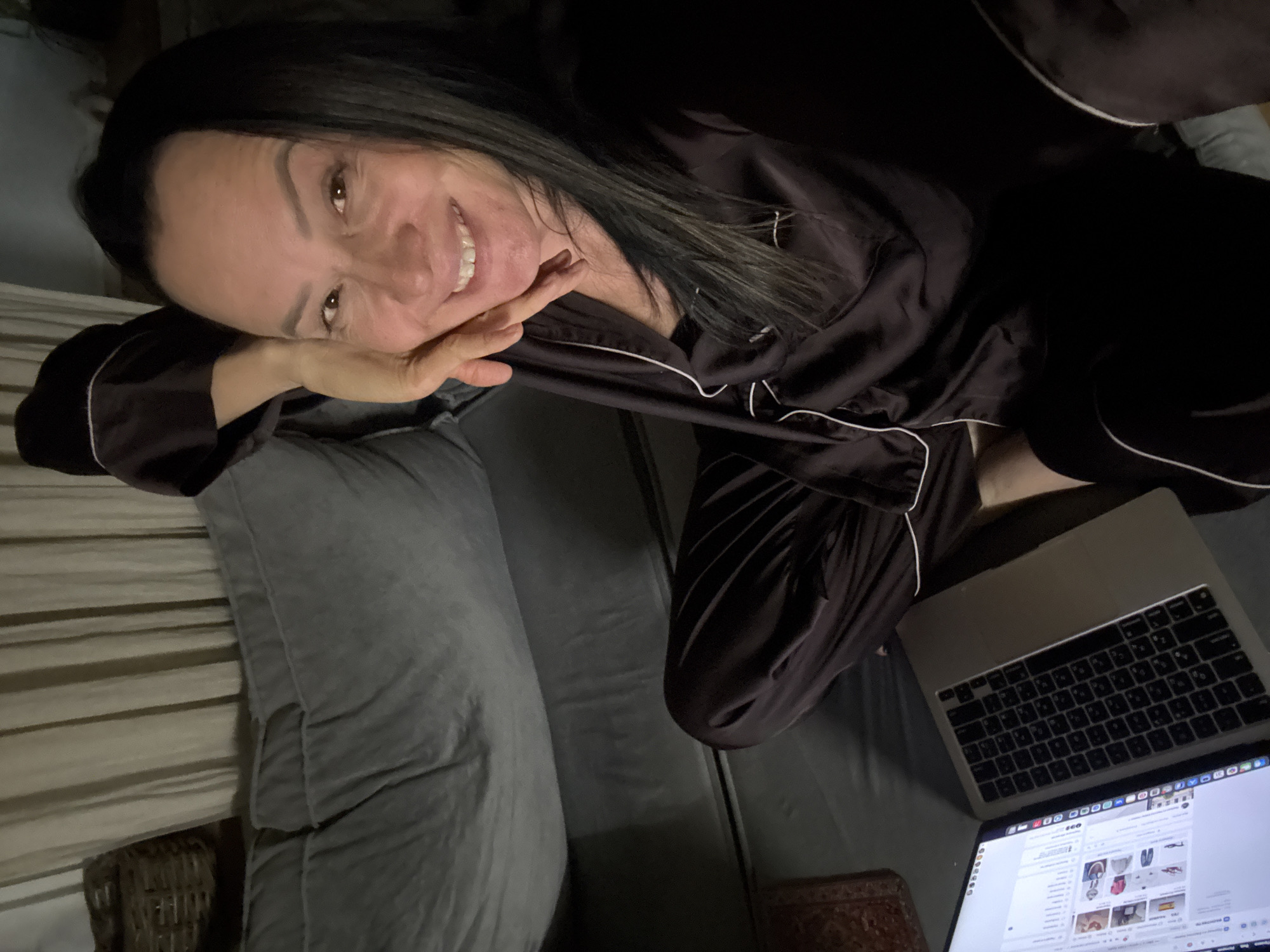 A woman is lying on a sofa while using a laptop. She is wearing dark pajamas with white trim. Her head is resting on her hand, and she is smiling at the camera. The laptop screen is visible, displaying what appears to be a website with product images. The sofa is gray, and a pillow is behind the woman. The location is Chaikovsky, Russia.