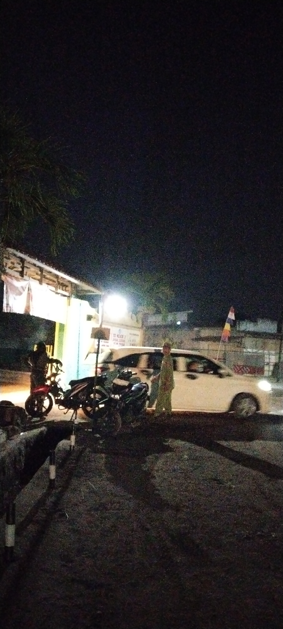This night image captures a street scene in Sido Rahayu, Indonesia, featuring a white sedan, several motorcycles, and two individuals. The sedan is parked on the right side of the road, facing away from the viewer. To the left of the sedan, two motorcycles are parked. One appears to be a dark-colored scooter, and the other is a lighter-colored motorcycle.
Two individuals are visible in the scene. One person, wearing a light-colored robe, stands next to the white sedan on the right. Another individual, wearing dark clothing, is standing next to a motorcycle on the left. The area is illuminated by artificial lighting, casting shadows on the asphalt road. In the background, a building with awnings and a sign displaying text is visible, along with some trees. A small flag with three horizontal bands of color is attached to the white sedan. The overall environment suggests a residential or commercial street at night.
