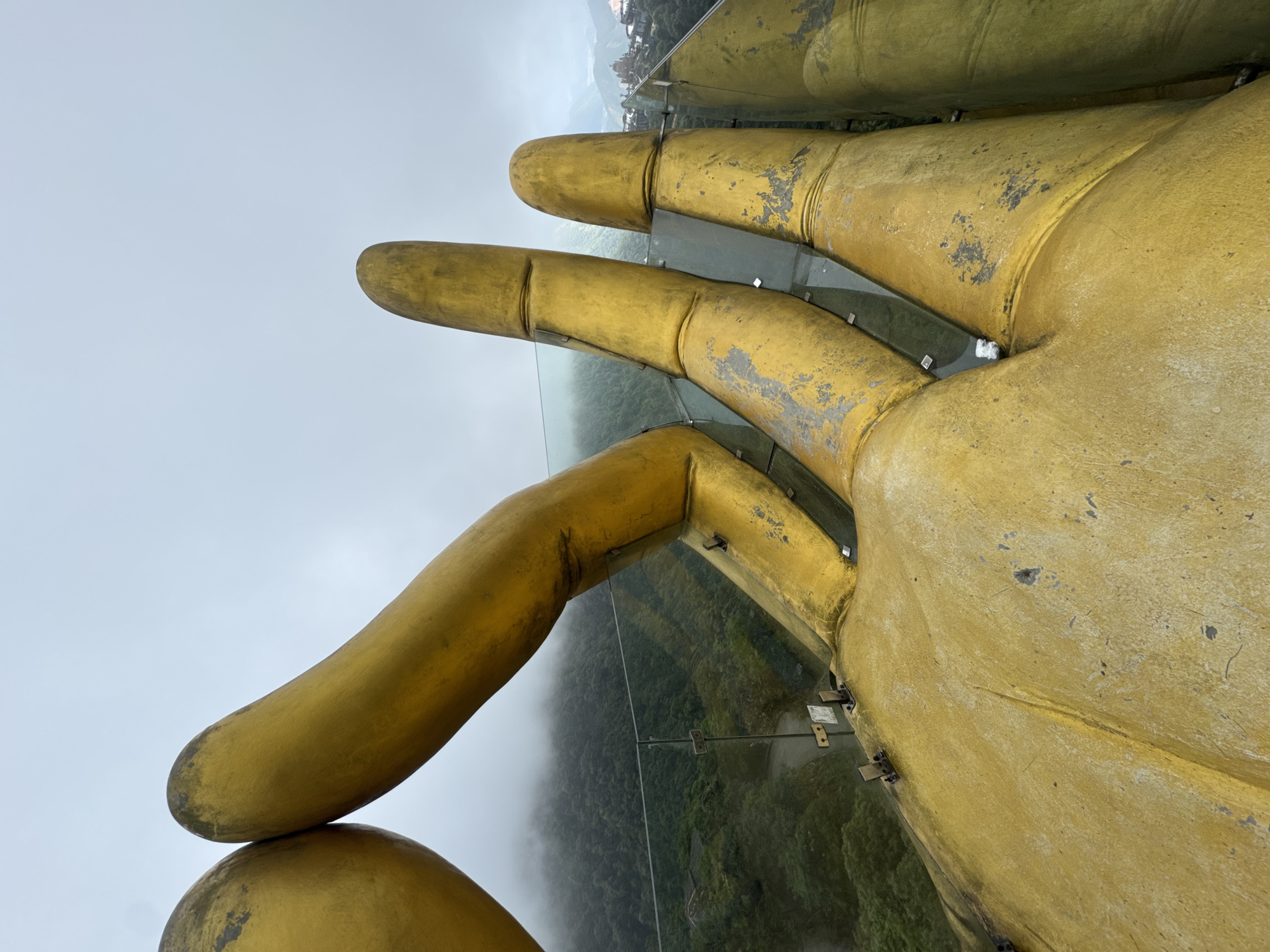 The image depicts a view from the Golden Bridge in Binh Lu, Vietnam, focusing on the massive golden hands supporting the bridge. The perspective is from the bridge looking out, showing the fingers of the hand, the glass walkway, and the forested landscape below, which is partially obscured by mist. The golden paint on the hand shows signs of wear and age. The sky is overcast.