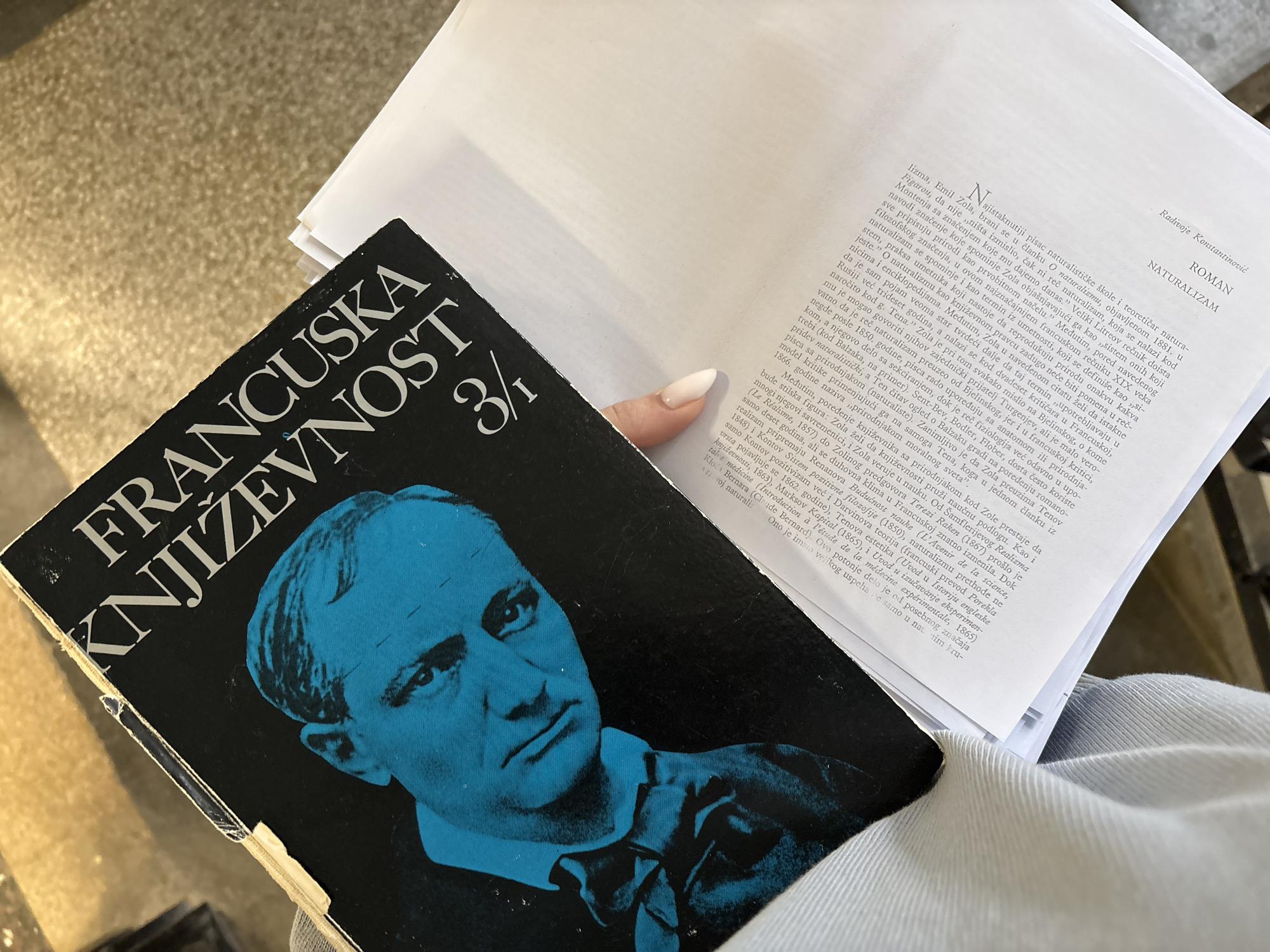 The image shows a person holding a book titled "Francuska Knjizevnost 3/1" (French Literature 3/1) in their hand. The book has a black cover with a blue portrait of a man. Next to the book is a stack of papers with text written in a Cyrillic script. The Cyrillic text appears to be a chapter of a book titled "ROMAN" and "NATURALIZAM". The location in the city of Kragujevac, Serbia, is unknown.