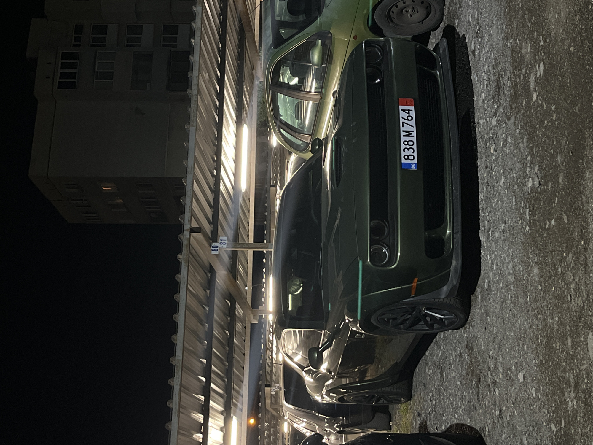 The image shows a dark green Dodge Challenger parked under a metal canopy in an outdoor parking area, likely at night. Next to it is a lighter green compact car, partially visible. The Dodge Challenger has a Bulgarian license plate "838 M 764." In the background, there is a tall, residential building with multiple windows. Based on the architectural style and the presence of Bulgarian license plates, the location is likely in Sofia, Bulgaria.