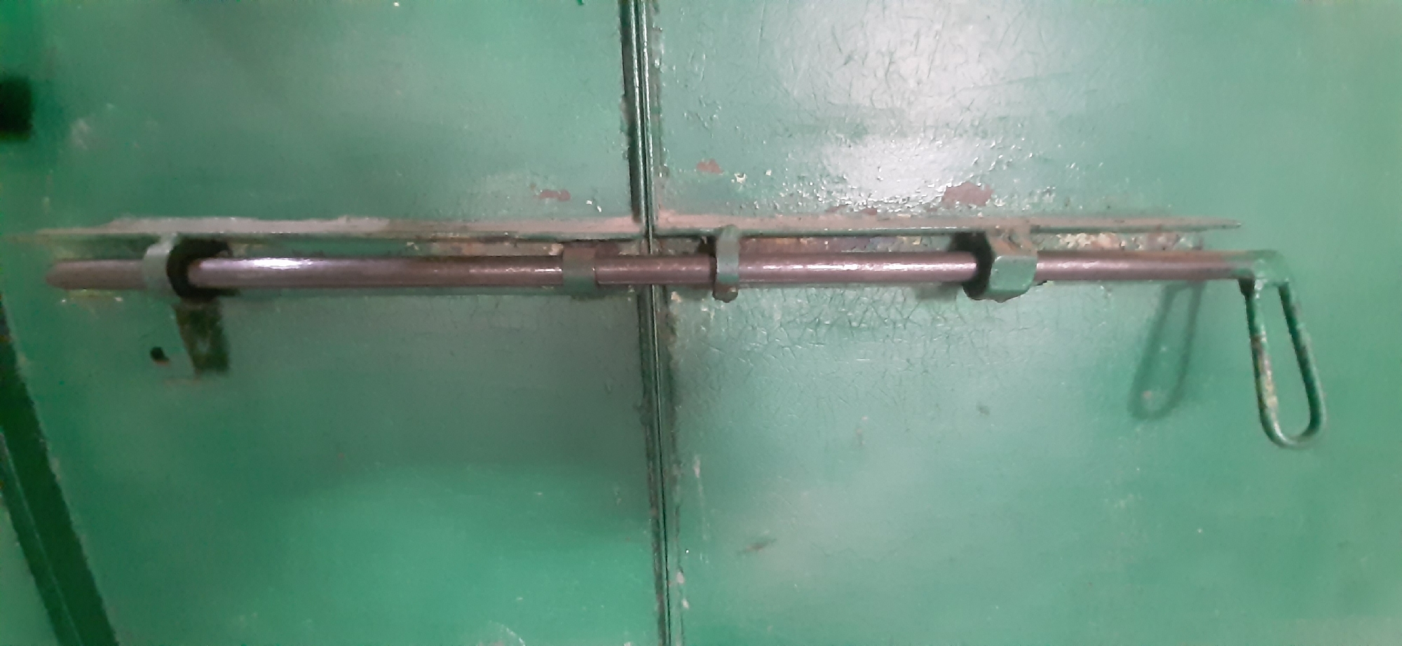 The image shows a close-up of a green, metal door with a sliding bolt latch. The door appears to be old, with peeling paint and visible wear and tear. The latch is made of metal and is in a locked position, with the bolt extended across the seam of the door. The background is a solid green color, with some texture from the paint. There are no people or other objects visible in the frame. The lighting suggests it could be indoors or outdoors on an overcast day, as there are no strong shadows. The setting is likely utilitarian, given the nature of the door and latch. There is no visible text in the image. The overall impression is one of security or a closed-off space.