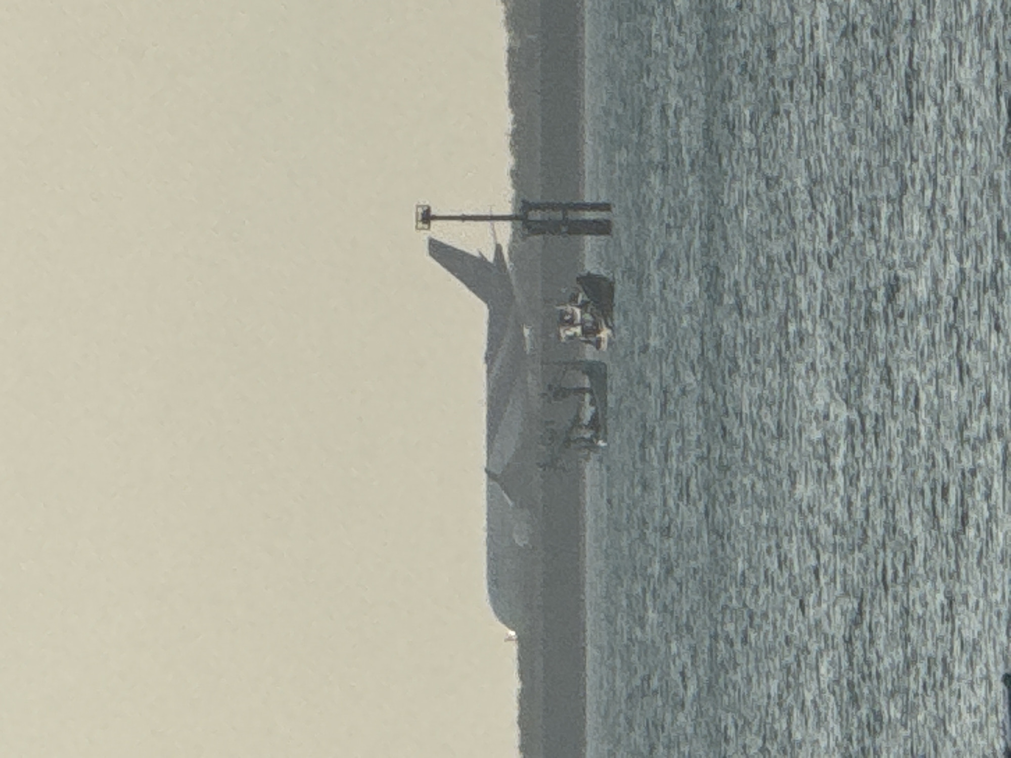 A silhouetted airplane is seen in the hazy distance, its outline sharp against the pale sky, as it appears to be either taking off or landing on a runway that stretches horizontally across the frame. In the foreground, the surface of the water is textured with ripples, suggesting a gentle movement. Two small boats are visible on the water, one with a more defined structure and several figures on board, while the other is less distinct. A tall, slender marker post, possibly a navigation buoy or a channel marker, stands vertically in the water between the two boats. The overall atmosphere is subdued, with muted colors and a sense of distance, hinting at a coastal or marine environment.