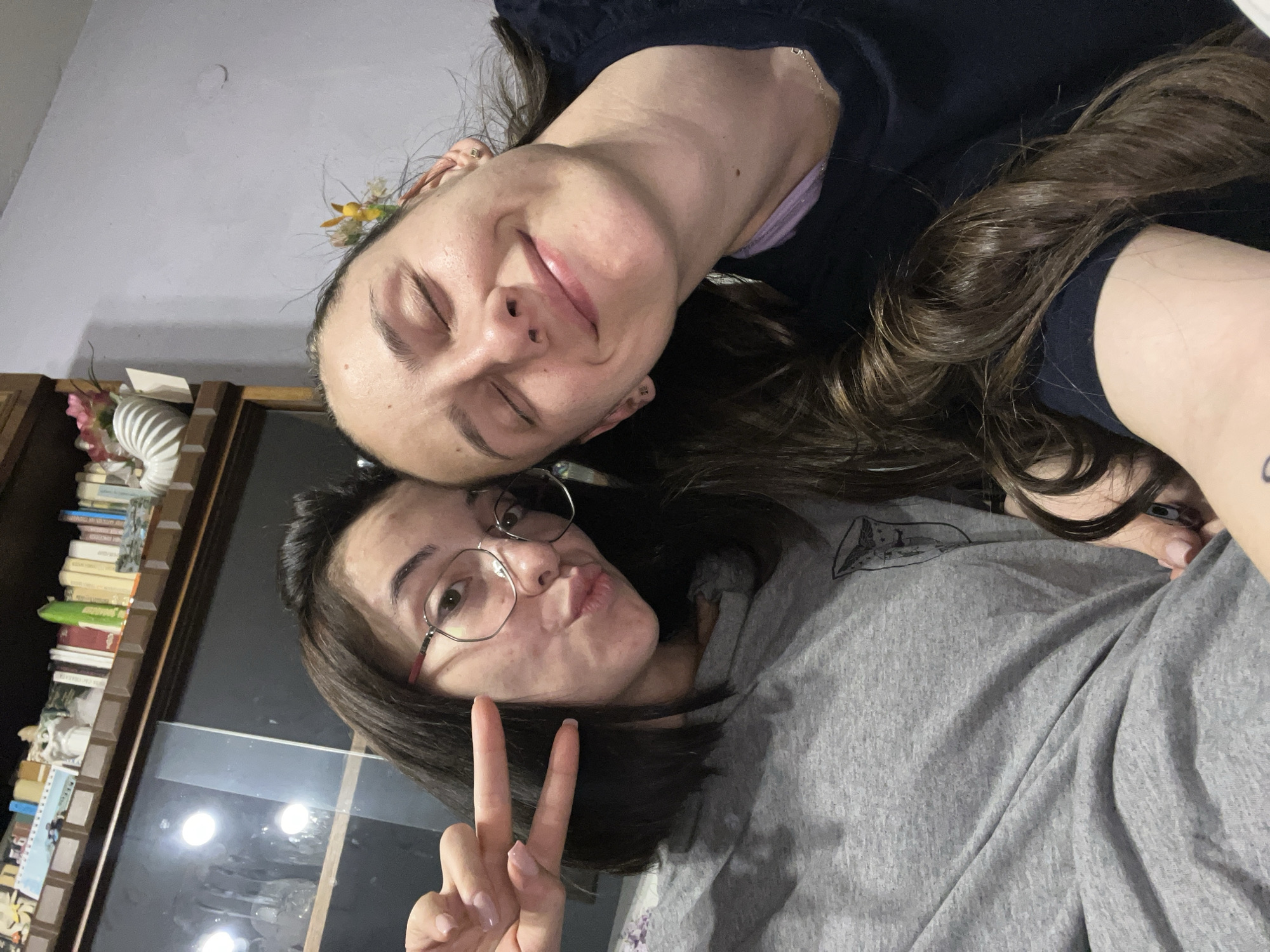 Two women are posing closely together. The woman on top is lying with her eyes closed and smiling slightly. She has brown hair and is wearing a dark blue shirt. The woman below is wearing glasses, pouting with her lips, and giving the peace sign. She has shoulder-length brown hair and is wearing a gray sweatshirt. They are in an interior setting with a bookcase visible in the background. No information to indicate location in Pamukchii, Bulgaria.