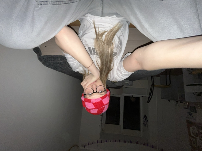 The image displays a person, likely a young individual, in an inverted orientation, suggesting they are lying down with their legs elevated or the image has been rotated 90 degrees clockwise. The perspective is a close-up selfie taken from a low angle.
The central figure has long, light-colored hair (blonde or light brown) that cascades downwards towards the upper part of the frame, in line with the inverted pose. They are wearing a white, short-sleeved t-shirt, on which a dark, block-lettered design or text is partially visible on the right side of the chest, though not clearly legible. The person's legs, clad in light grey or faded blue jeans or sweatpants, are prominent in the foreground, bent at the knees, framing the view.
The individual's face is partially obscured by their hands, which are held close to the face. One hand covers part of the mouth and chin, while the other rests near the cheek. They wear round-rimmed glasses with dark frames. A distinctive red and pink checkered (or tartan-like) pattern headband or ear warmer is worn across the forehead, covering the eyebrows. Only the eyes and a portion of the nose are clearly visible above the hands, making a precise emotional expression difficult to discern, but the overall impression is casual and relaxed. A thin, light-colored bracelet is visible on one wrist.
The background suggests an indoor setting, likely a bedroom. Behind the person's head, there is a bed or sofa covered with a dark grey or black textured blanket or throw. A wooden headboard or furniture frame is visible directly above this. To the right side of the frame, a window with dark panes indicates it is either dark outside or the blinds are closed. Below the window, a light-colored surface, possibly a windowsill or part of the wall, is visible. Further to the right, some dimly lit furniture or decorative items, along with a glimpse of patterned wallpaper or a wall hanging, can be seen. The overall lighting is dim, suggesting it is evening or night. There is no visible, clearly legible text within the image apart from the obscured design on the t-shirt.