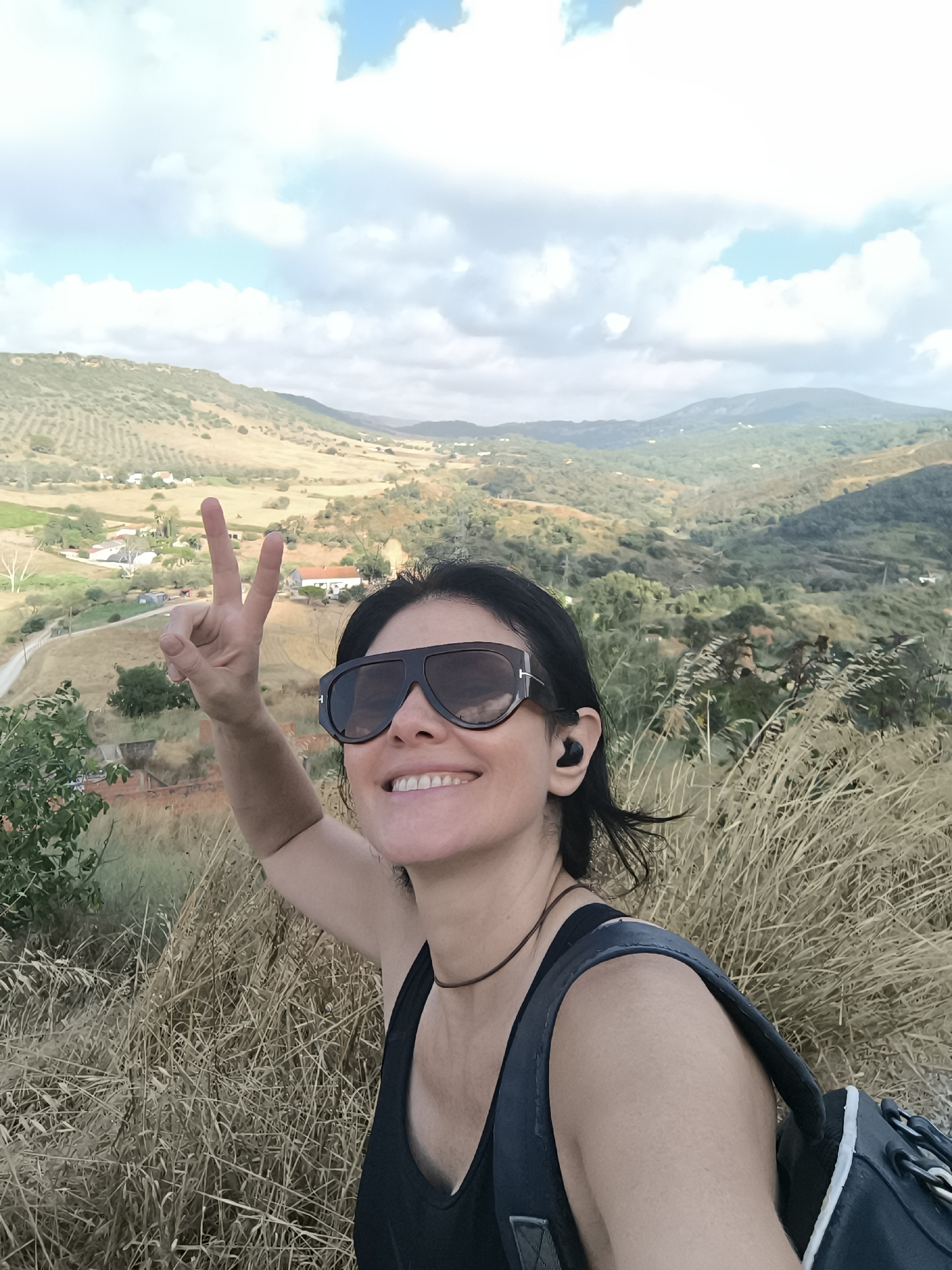 A woman with dark hair, sunglasses, and a black tank top is taking a selfie in a rural, hilly landscape. She is smiling and holding up two fingers in a peace sign. The background shows rolling hills covered in vegetation, scattered houses, and a cloudy sky. She is wearing black wireless earbuds. The location is identified as Palmela, Portugal. The foreground is filled with dry, tall grass, and she is wearing a backpack.