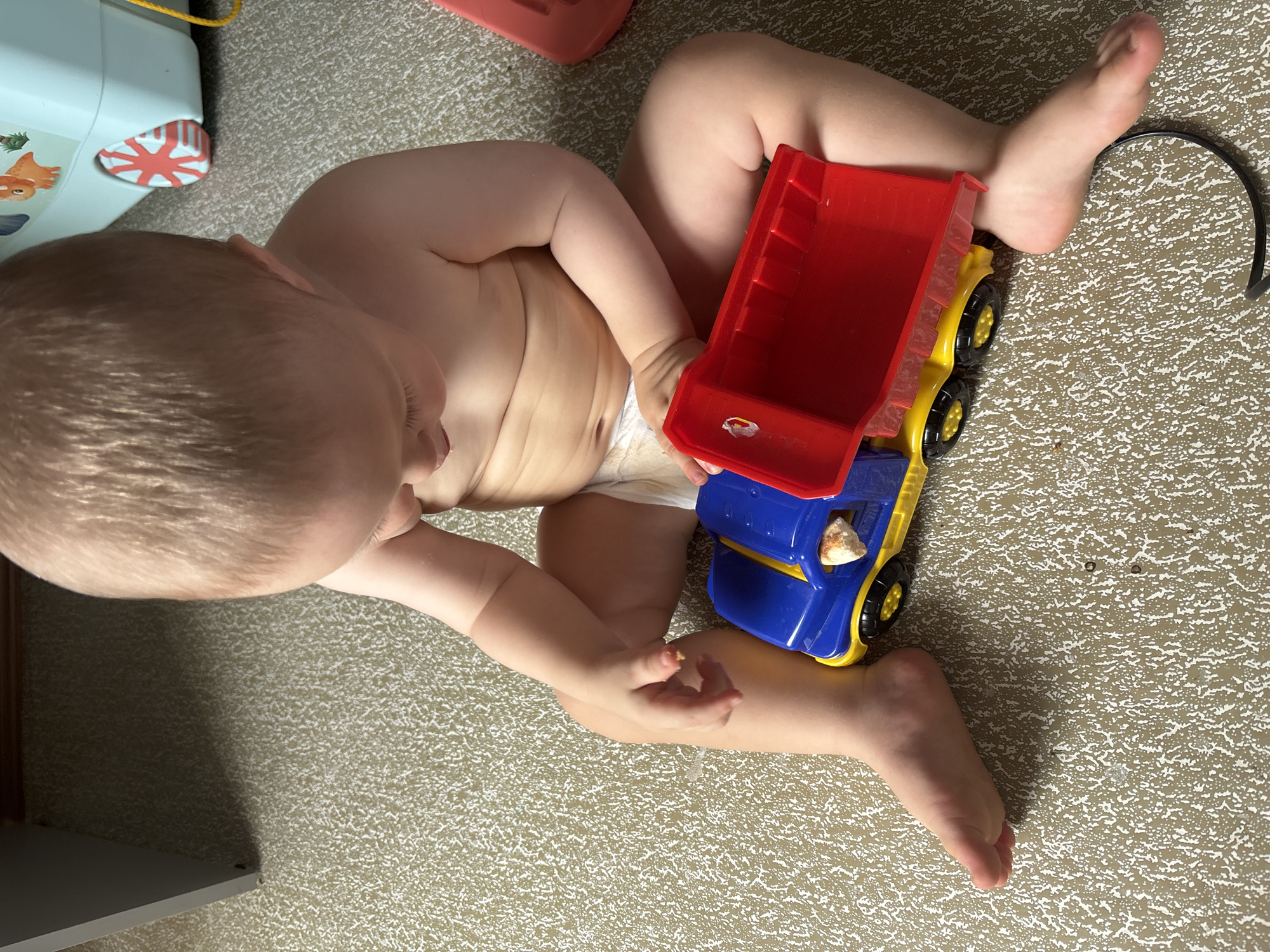 A baby is sitting on a textured floor, playing with a toy dump truck. The baby is wearing a diaper and is looking down at the toy. The dump truck is blue and red with yellow wheels and has a piece of food inside the truck bed. The environment is likely indoors, given the presence of a toy and the flooring. There is no information to suggest the location is in Naberezhnye Chelny, Russia.