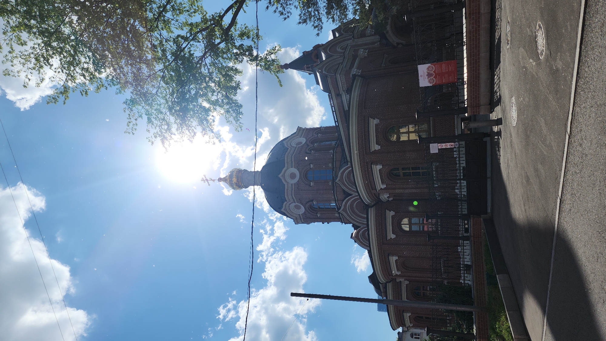 The image, rotated clockwise, captures a large, ornate red-brick Orthodox church or cathedral under a bright, clear sky. The building features intricate architectural details, including rounded windows with arched tops and decorative light-colored trim. A prominent dark dome, topped with a golden, onion-shaped cupola and an Orthodox cross, stands out against the blue sky, reflecting the sunlight. Another smaller dome or spire is visible towards the right side of the building.
The sky is a brilliant blue with scattered white, fluffy clouds. The sun is visible in the upper left, casting a strong lens flare and bright rays, suggesting midday or early afternoon. Green tree foliage is visible at the top, framing the sky.
A black metal fence runs along the perimeter of the building, separating it from an asphalt street or sidewalk. Three circular manhole covers are visible on the street, which is bordered by a light gray curb. Thin black wires are faintly visible stretching across the sky.
Several signs are attached to the fence. A red rectangular sign displays circular emblems and unreadable text, likely in Cyrillic. Below it, a smaller white sign shows universal "no" symbols, possibly indicating restrictions like "no smoking" or "no photography." No people or specific events are depicted, presenting a tranquil scene of the architecture bathed in sunlight, consistent with the given location context of Moscow, Russia.