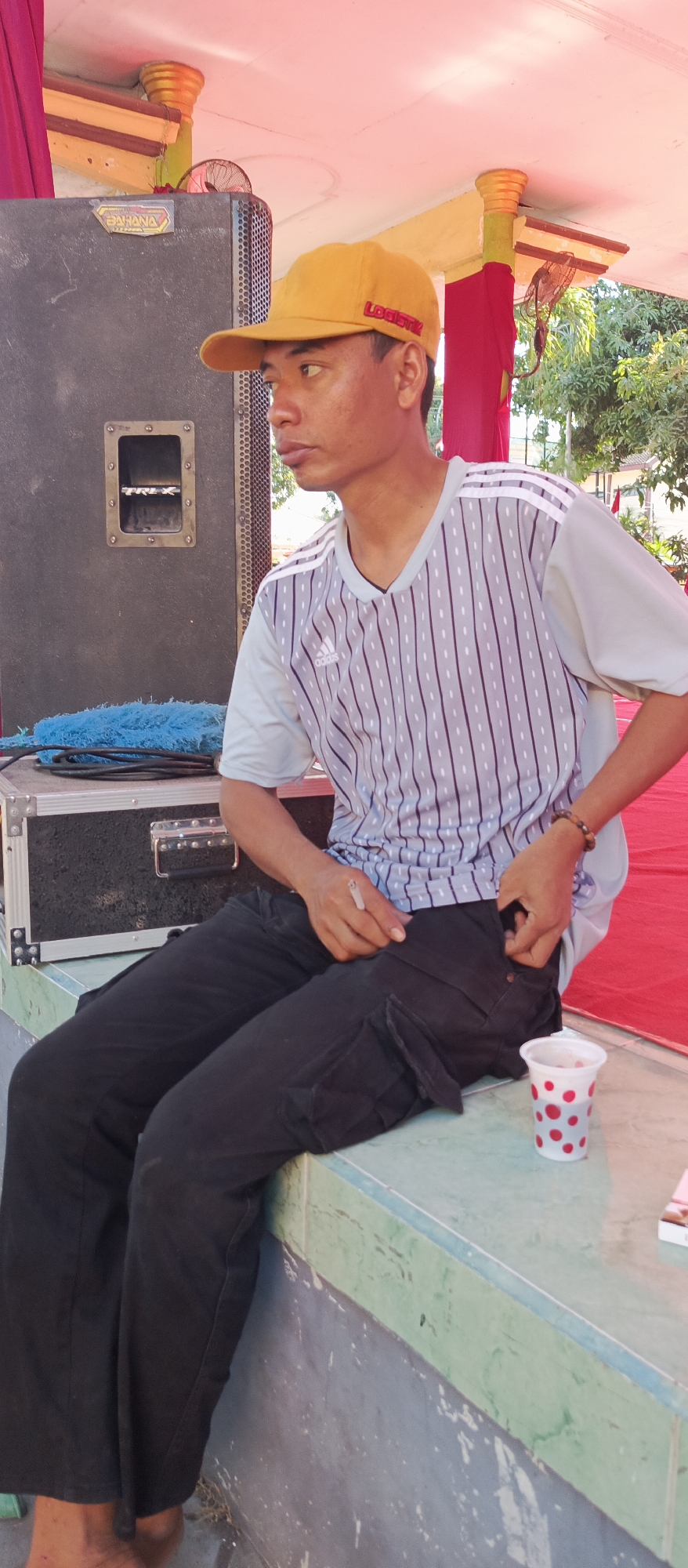 A young man wearing a yellow baseball cap and a grey and white striped t-shirt sits on a ledge in Arjasa, Indonesia. He is wearing black cargo pants and is holding a cigarette in his right hand. A red and white polka-dotted cup sits on the ledge next to him. In the background, there is a large black speaker and a red tablecloth. The sky is visible in the upper part of the image and appears to be overcast. The overall scene is informal and appears to be outdoors.