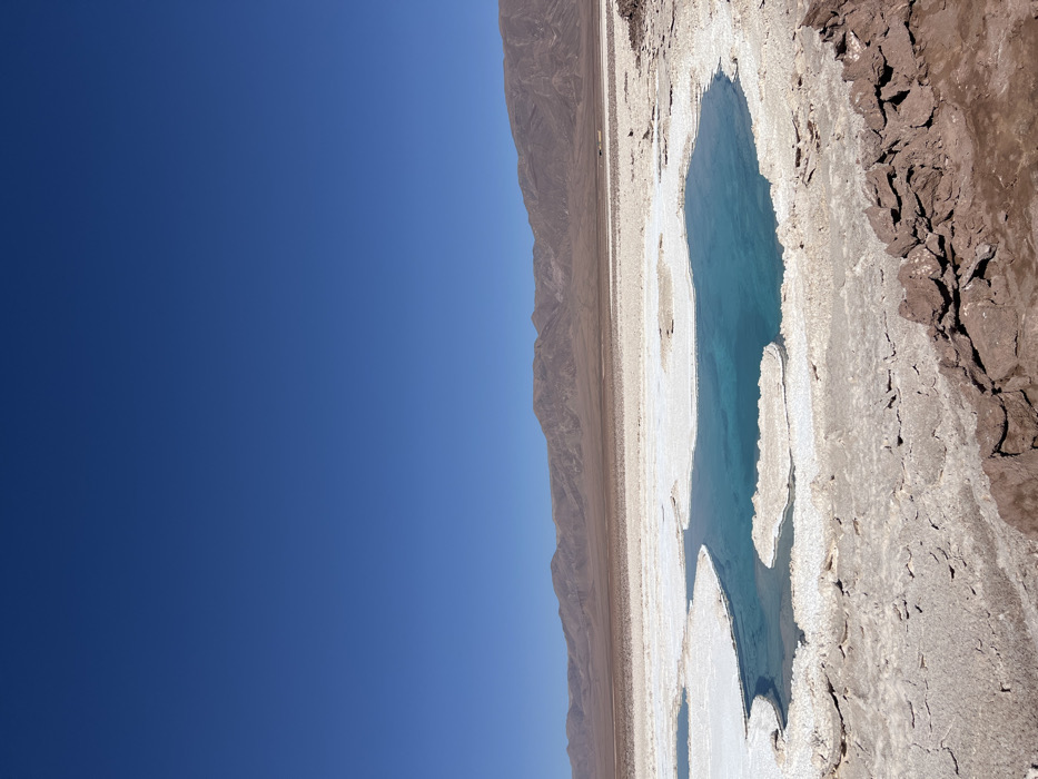 A landscape features a clear blue sky contrasting with a terrain of mountains and a unique salt flat formation. A body of turquoise water, irregularly shaped, sits amidst the cracked and textured earth, bordered by a white crust. The mountains appear barren, stretching along the horizon. The composition presents a stark, arid environment with a vibrant splash of color in the water.
