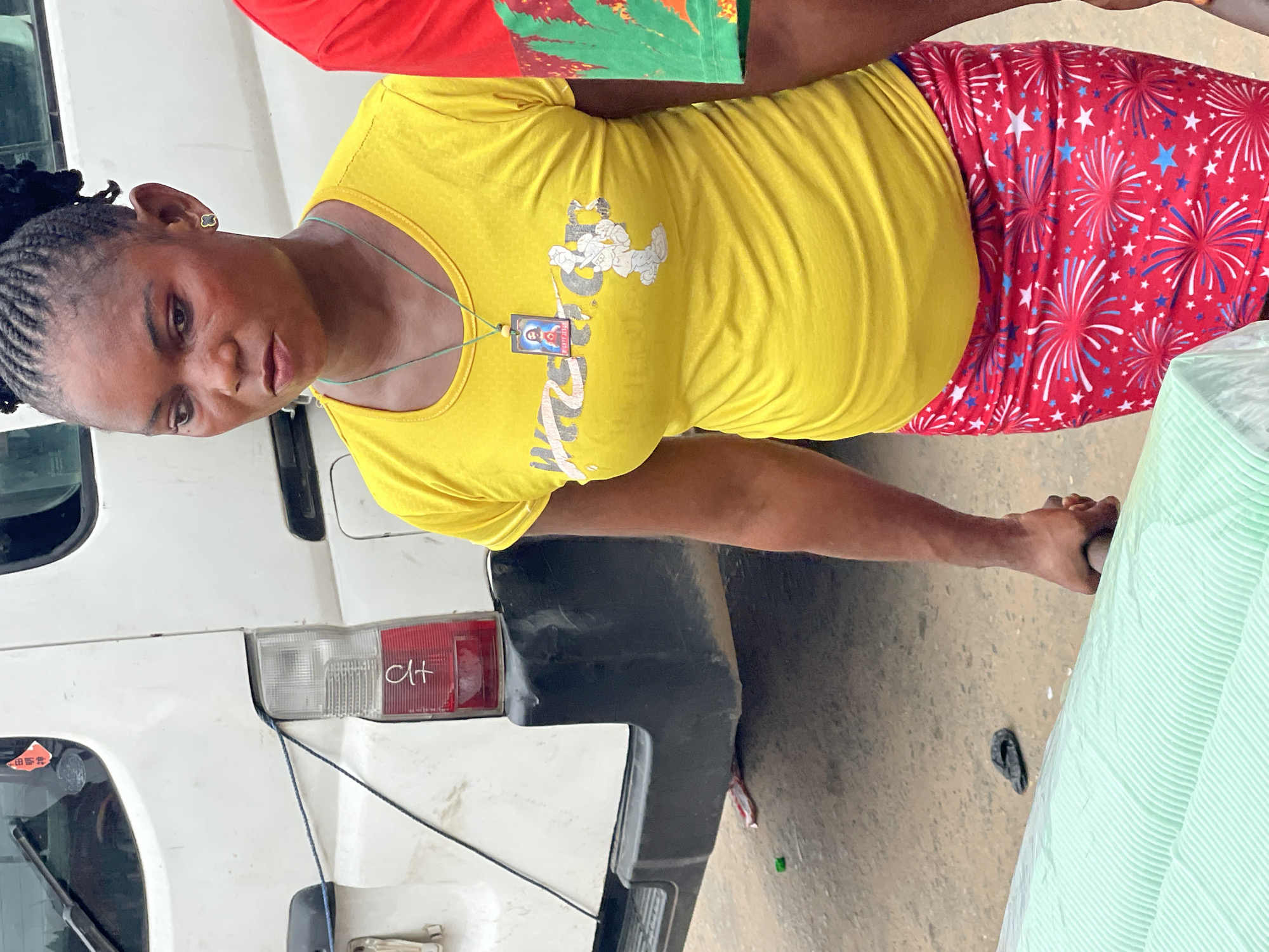 A woman with braided hair stands in front of a white vehicle with the letters "CH" written on the taillight. She wears a yellow t-shirt, patterned red and blue pants, and a pendant necklace. Her expression is neutral. She is holding a wrapped green item. The location appears to be outdoors, possibly in the city of Onitsha, Nigeria, based on cultural elements evident in her dress, hairstyle, and the vehicle.