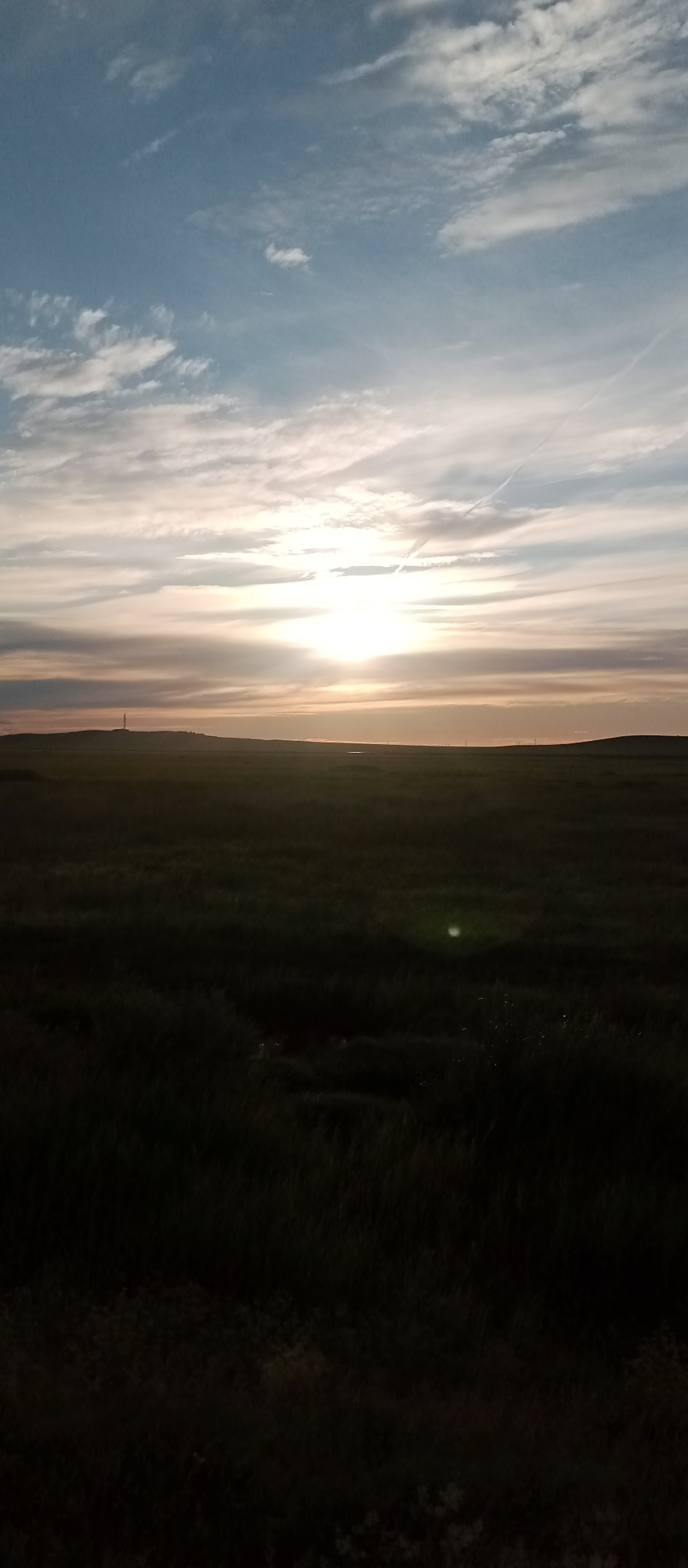 The image shows a wide, flat landscape under a cloudy sky during sunset. The sun is low on the horizon, casting a warm glow and creating streaks of light across the sky. The foreground is a dark, grassy field, with some areas appearing almost black due to the low light. In the distance, a low hill is visible, with a tall, slender structure on top, possibly a radio tower or pylon. The horizon line is sharp and distinct, separating the land from the sky. The overall impression is one of open space and natural beauty, with the sunset adding a dramatic and serene atmosphere. The location is identified as Marinovka, Kazakhstan.