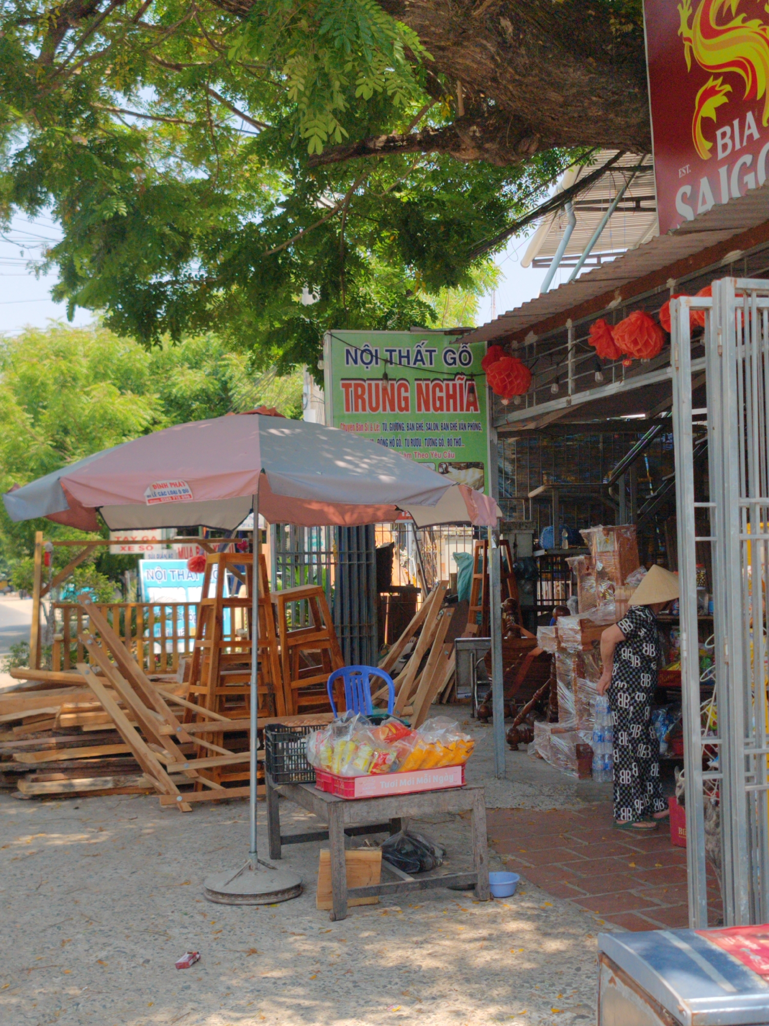 The image depicts an outdoor furniture store in Khánh Hải, Vietnam. Various wooden planks, ladders, and furniture frames are visible in front of the store, partially shaded by a large umbrella. A sign reading "NỘI THẤT GỖ TRUNG NGHĨA" is displayed prominently. A woman wearing a traditional conical hat stands near the store's entrance, amidst furniture items wrapped in plastic. Red lanterns hang from the store's roof. A "Bia Saigon" sign is also partially visible on the right side of the image. A table with a basket of packaged goods is placed in front of the store.