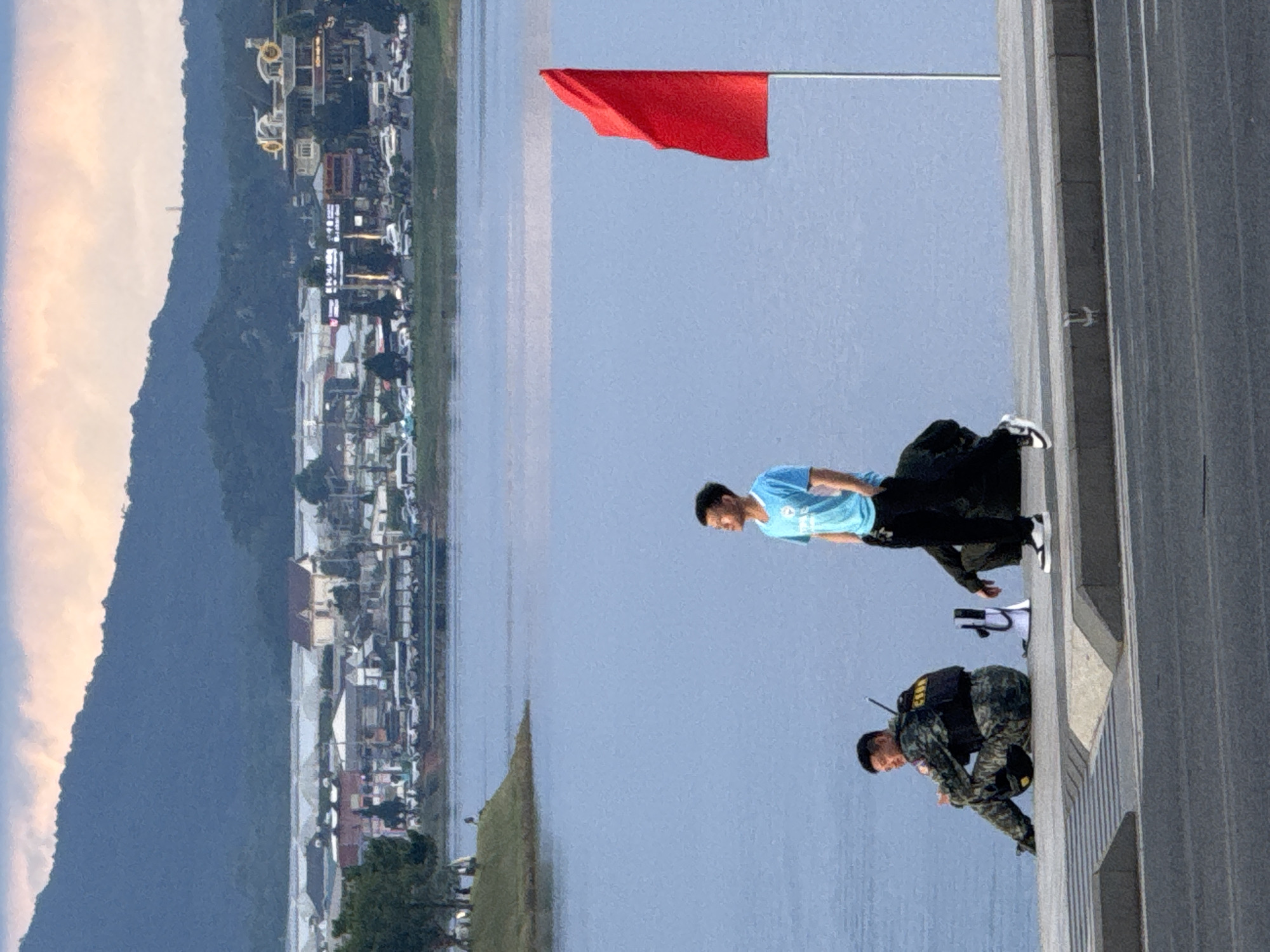 The image captures a serene outdoor scene, likely in the city of Da Lat, Vietnam, given the lush green landscape and the distinctive architecture visible in the background. In the foreground, a man in a blue t-shirt and black pants stands by a calm body of water, looking towards the sky. To his left, another man, dressed in a military-style uniform with a vest and tactical pants, is seated on a ledge overlooking the water. A red flag flutters on a pole to the right of the standing man, adding a splash of color to the scene. The background reveals a sprawling cityscape with buildings and mountains stretching into the distance, hinting at a blend of urban life and natural beauty. The overall impression is one of tranquility and observation.