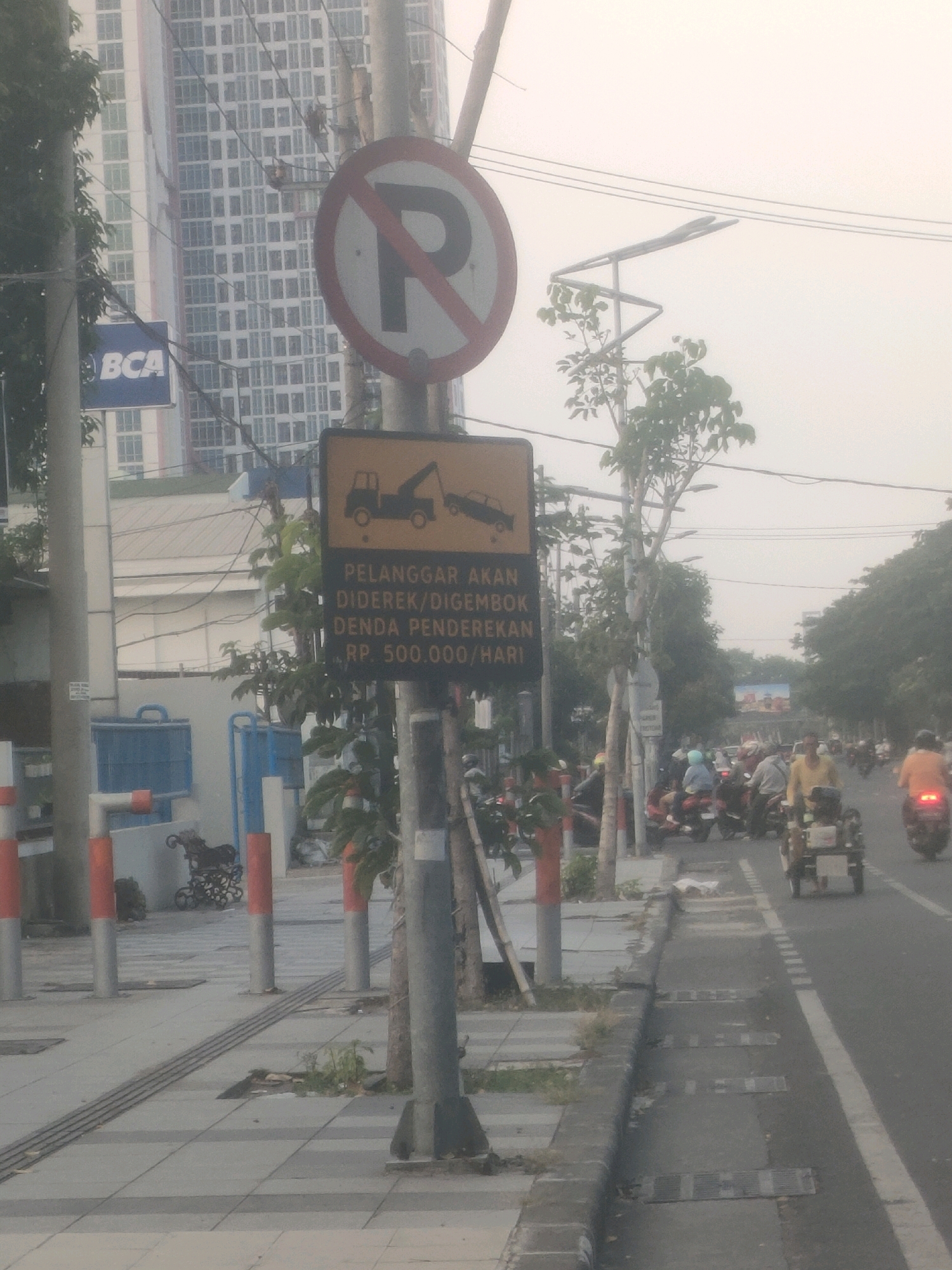 The image depicts a street scene in Surabaya, Indonesia, likely a commercial or residential area given the presence of a tall building. A prominent "no parking" sign with a red circle and diagonal slash over a 'P' is visible on a pole. Adjacent to it is a yellow rectangular sign with an illustration of a tow truck hooking up a car, and text below indicating that violators will be towed and fined 500,000 Indonesian Rupiah per day. The street is lined with a sidewalk featuring decorative bollards and trees. In the background, a modern high-rise building with a grid-like facade stands out. The road is busy with traffic, primarily motorcycles, and several individuals are seen riding them. The overall atmosphere suggests an urban environment during daytime, possibly with moderate weather conditions due to the hazy sky.