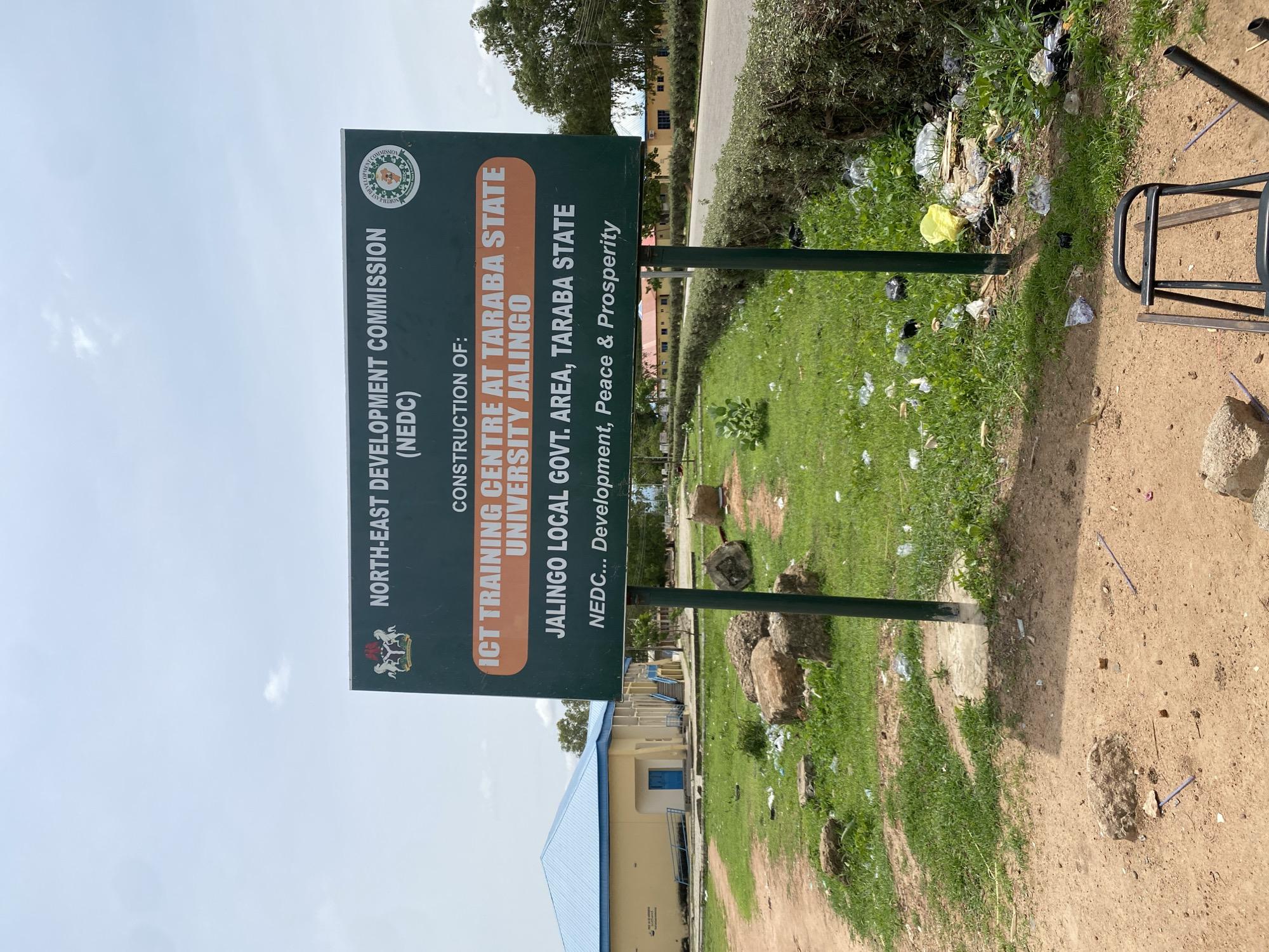 A green billboard stands in front of a yellow building with a blue roof, in Jalingo, Nigeria. The billboard announces the construction of an ICT Training Centre at Taraba State University, Jalingo, by the North-East Development Commission (NEDC). The area surrounding the billboard is unkempt, with scattered trash and some rocks on the ground. A broken chair lies on the dirt in the foreground. The sky is overcast.