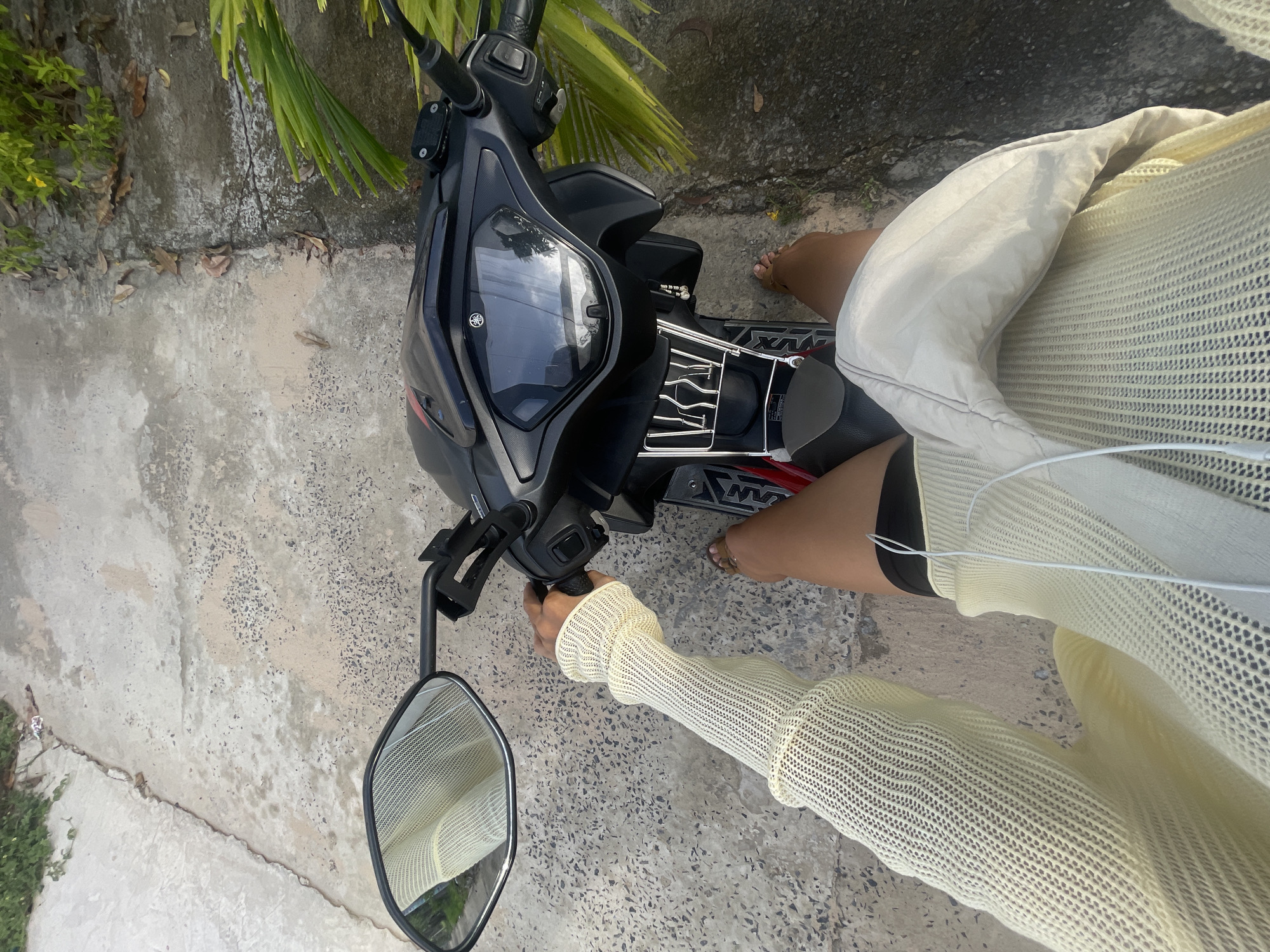 The image, taken in Phu Thuy Ward, Vietnam, is rotated 90 degrees clockwise and shows a person sitting on or next to a black and red Yamaha scooter on a bright, sunny day. The person, with fair skin and bare feet wearing sandals, is wearing a light yellow, sheer, long-sleeved top or sweater and dark shorts. A light-colored, possibly padded, bag rests on their lap or the scooter seat, and white wired earphones are visible.
The person's right hand is on the scooter's handlebar. The scooter features a front storage basket made of metal grids, a dashboard with a visible "YAMAHA" logo, and a side mirror reflecting part of the person's attire and the sky. The text "NVX" is partially visible on the scooter's side panel.
The setting is outdoors, featuring a concrete or paved ground surface with scattered pebbles and cracks. Along one edge of the paved area, there is sparse greenery, including bushes and palm-like leaves, along with some dry leaves. The overall lighting suggests mid-day sunlight.