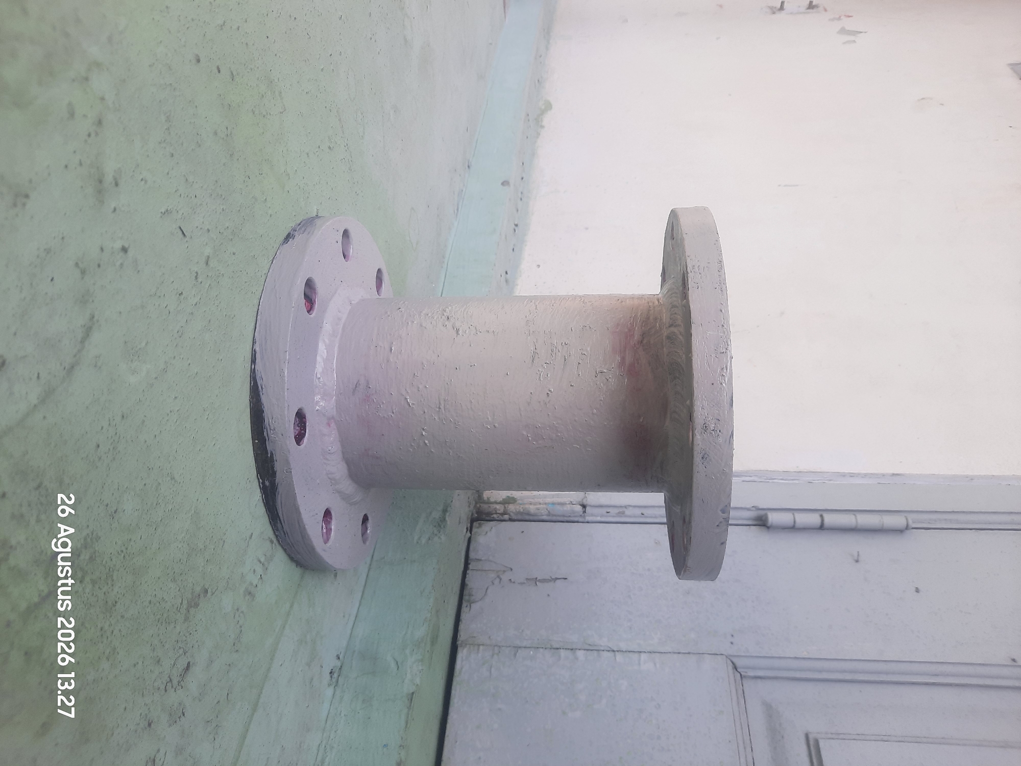 In Gending, Indonesia, a light gray pipe fitting stands on a light green concrete floor. The fitting has a cylindrical body with flanges at both the top and bottom. The top flange is circular, while the bottom flange is wider and features several bolt holes, some of which contain what appears to be red material. Behind the fitting, a light-colored wall transitions into a light blue door. The wall has some scuff marks and peeling paint. The floor shows some texture and scattered dark specks. A date stamp in the bottom right corner reads "26 Agustus 2026 13.27".