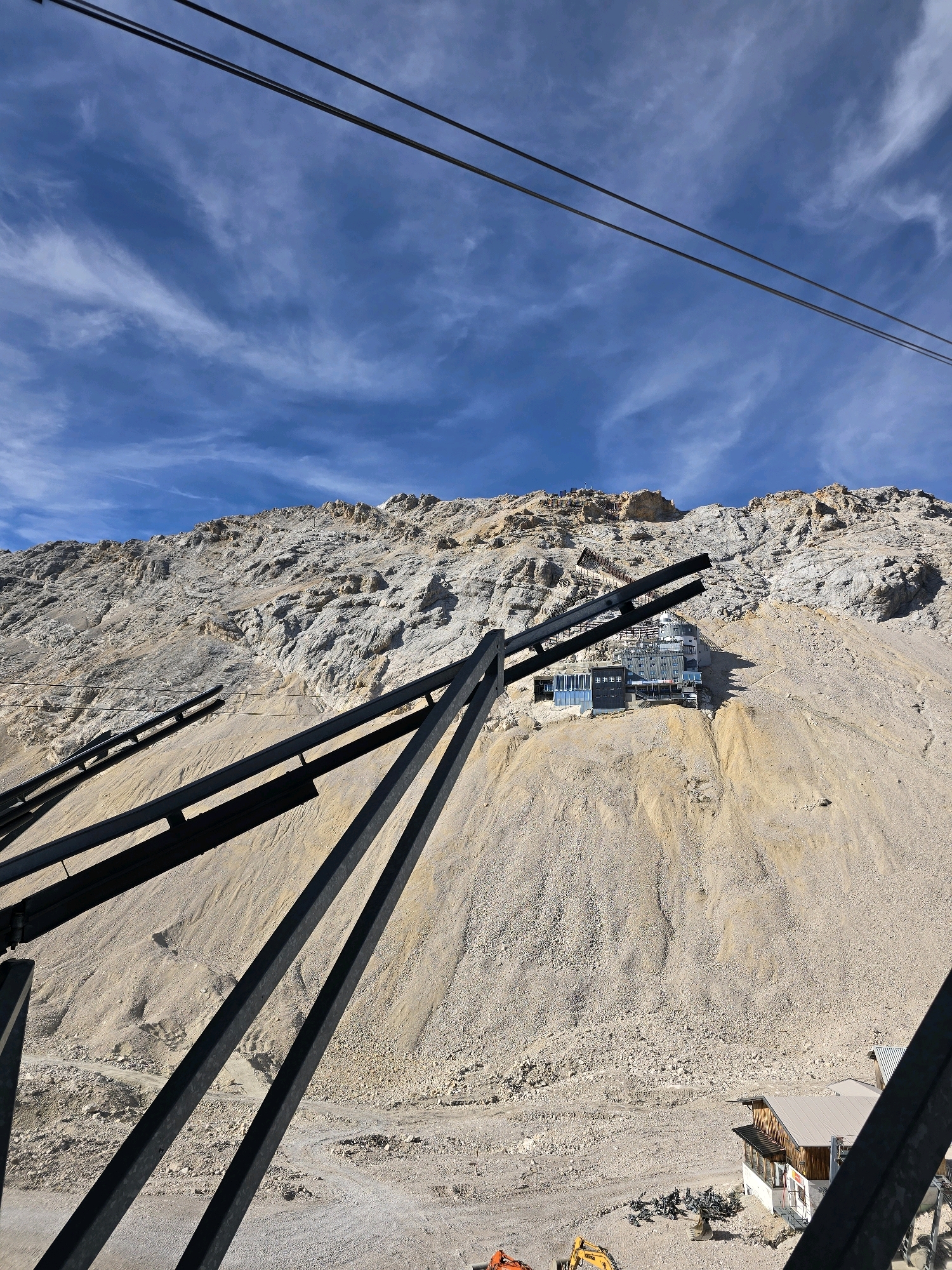 This is a vertical, high-angle, medium shot of a mountainside in Garmisch-Partenkirchen, Germany, dominated by a rocky, scree-covered slope under a bright blue sky with scattered cirrus clouds. In the foreground, dark metal beams, likely part of a cable car or infrastructure, extend diagonally across the frame. Further up the slope, a modern building with large glass windows is visible. At the base of the mountain, a small wooden building with a gray roof and a small structure next to it are present, alongside a yellow excavator and other construction materials. The overall scene depicts a rugged mountainous environment with ongoing construction or infrastructure development.