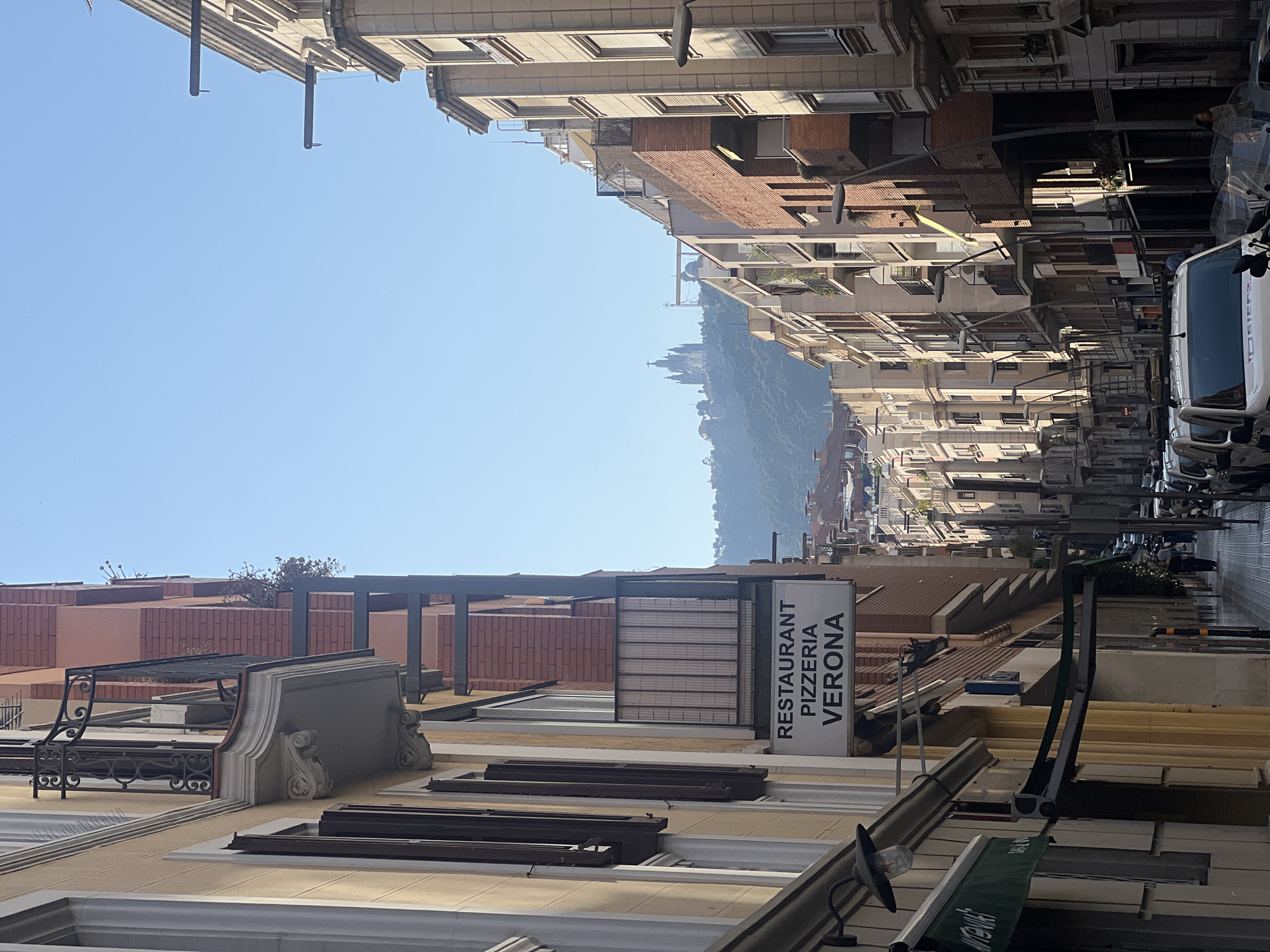 This is a vertical, eye-level, outdoor medium shot of a street scene in Barcelona, Spain. The sky is clear and blue, suggesting a sunny day. The buildings lining the street are tall and multi-story, with a mix of architectural styles, including ornate facades and more modern elements. The street itself appears to be a moderately busy thoroughfare with parked vehicles, including a white van.
On the right side of the street, a sign is prominently visible, displaying the text "RESTAURANT PIZZERIA VERONA" in large, black, sans-serif capital letters against a white background. This sign suggests a dining establishment is present. Further down the street, in the distance, a lush, green hillside is visible, topped with structures, possibly a historical site or monument, hinting at the city's landscape and heritage.
The overall impression is a vibrant urban environment bathed in sunlight, showcasing both the architectural character of Barcelona and its connection to the natural surroundings. The image captures a moment of everyday city life, with no specific event or activity being the central focus, but rather the atmosphere and composition of the urban landscape.