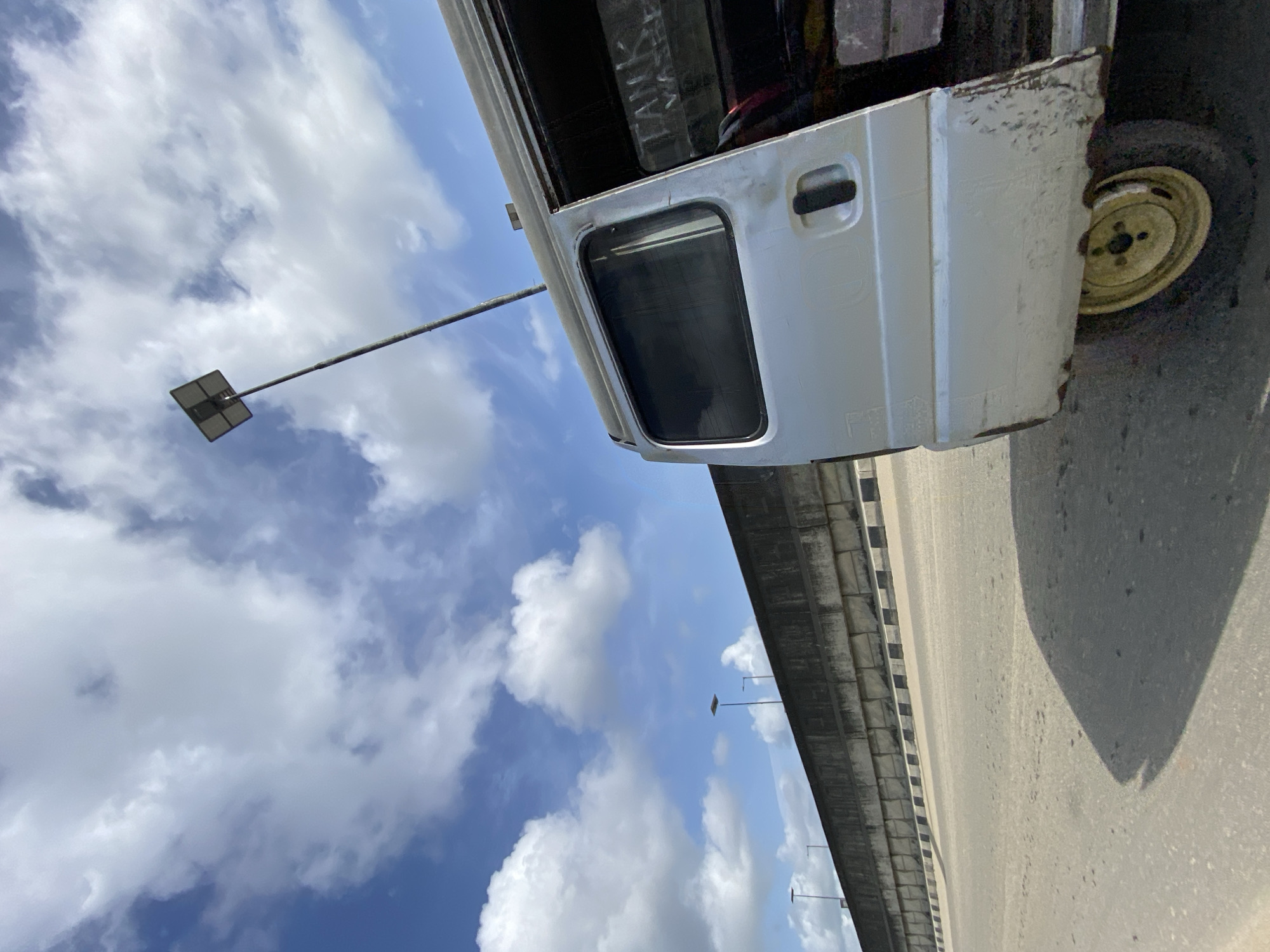 The image shows a white bus on a highway in Lekki, Nigeria. The bus is parked on the side of the road, and its door is open. The background of the image includes a blue sky with white clouds and a bridge. Streetlights are visible along the highway.