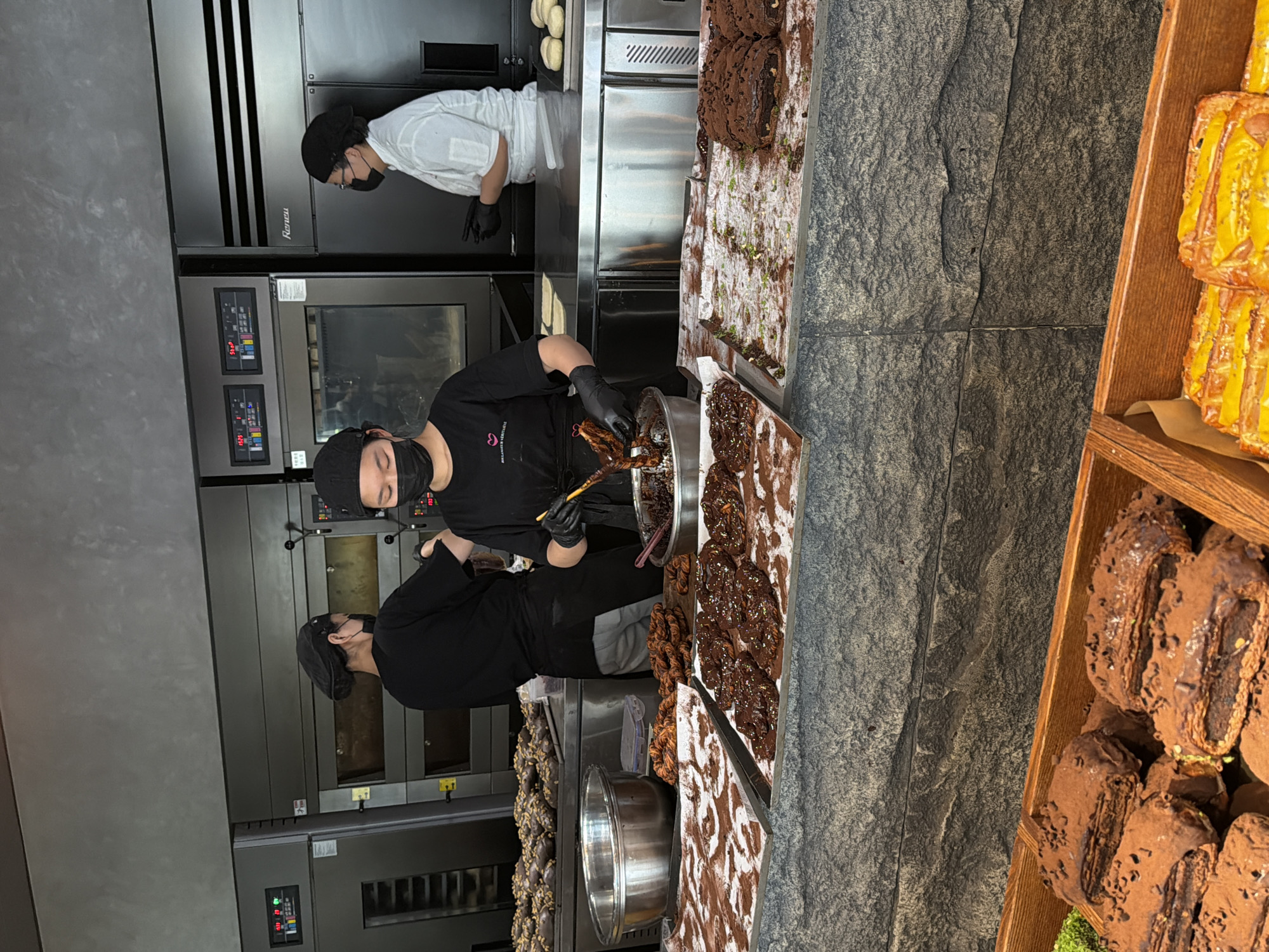 In a brightly lit bakery in Chongqing High-tech Zone, China, three individuals are working amidst bakery equipment and food displays. On the left, against a wall with multiple built-in ovens, two masked individuals wearing black uniforms, gloves, and head coverings, appear to be preparing food. One is actively mixing ingredients in a large metal bowl. To their right, a third individual, also masked, wears a white shirt and is positioned in front of a stainless-steel counter with trays of what appears to be pastries. Adjacent to the food preparation area is a dark stone-like surface, with trays displaying pastries with chocolate and possibly nuts. To the far right, there is a wooden tray containing pastries with a yellow topping.