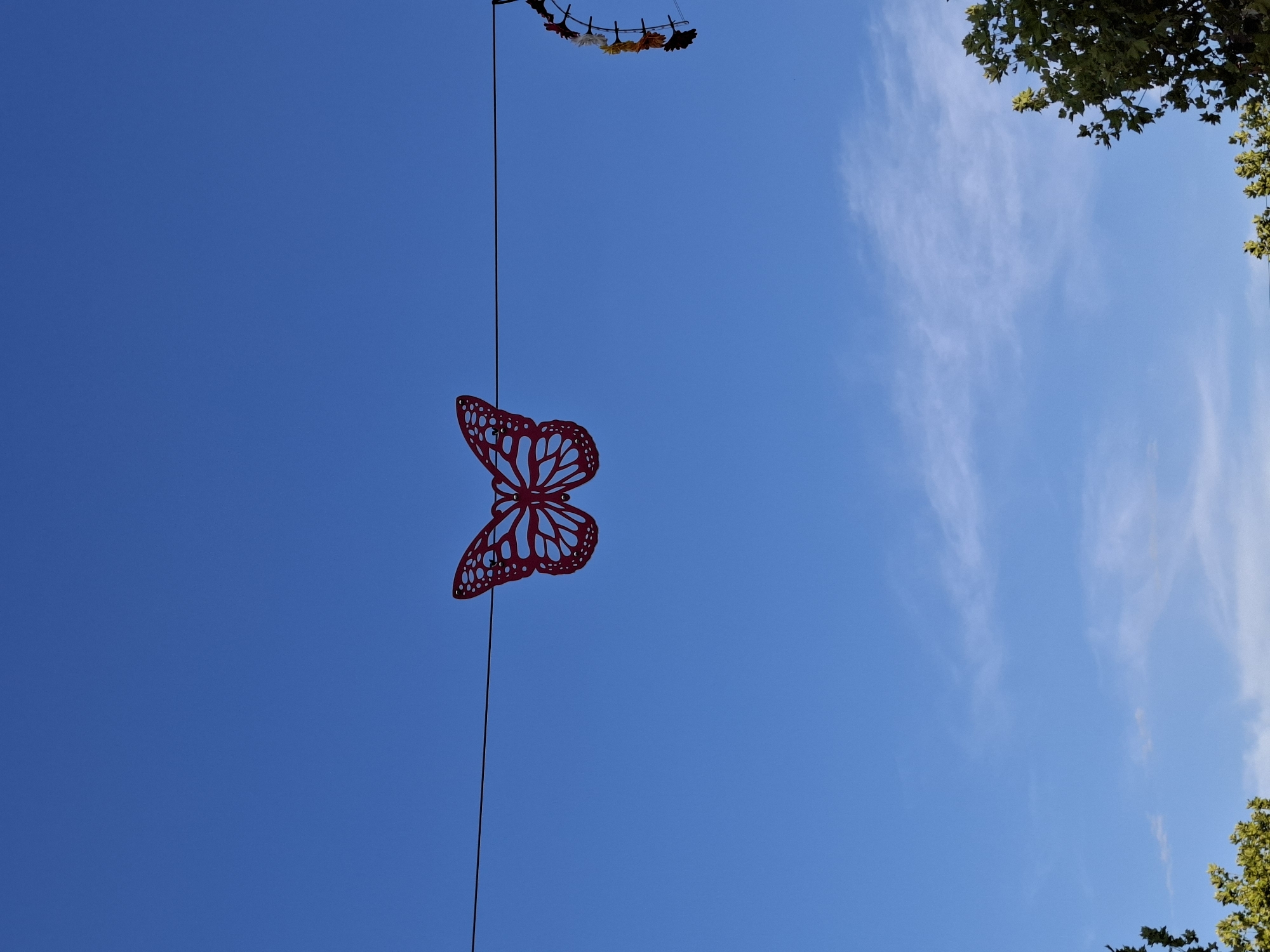 A red, cut-out butterfly decoration is suspended on a wire against a bright blue sky. The butterfly has intricate patterns within its wings. To the right of the butterfly, a portion of a decorative circular object with colorful flower-like elements is visible, also hanging from a wire. The bottom of the image shows the tops of green trees. The scene is likely outdoors, and the sky suggests it is daytime. The specific location in Rastatt, Germany, is not identifiable from the visual information provided in the image. There are no people or animals present.
