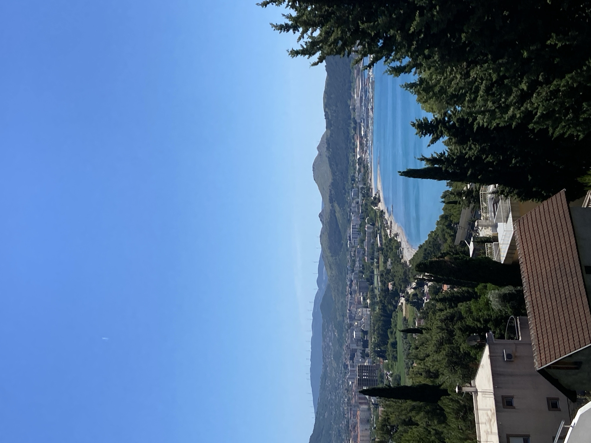 The image shows a panoramic view of Šušanj, Montenegro, extending from a foreground with rooftops and trees to a background of mountains, coastline, and the Adriatic Sea under a clear blue sky. The city is nestled along the coast and the mountains rise inland. There are buildings and vegetation interspersed throughout the landscape. A plane is visible in the sky. The image is shot from an elevated position.