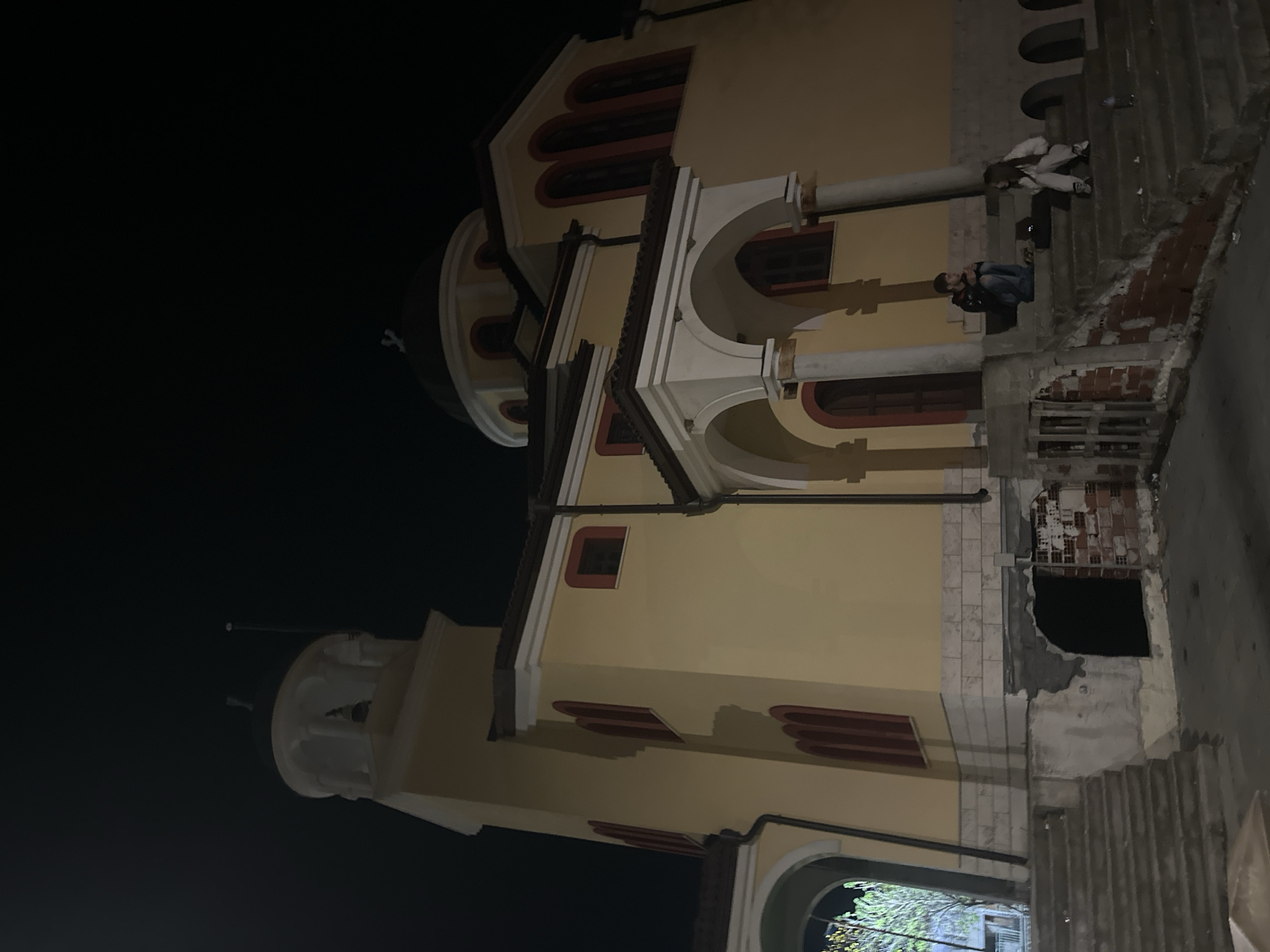 At night in Katuntsi, Bulgaria, a three-story yellow building stands with dark red window trim and arches. Two people sit on a concrete structure abutting the building. One person sits on the structure, facing the building. Another person, wearing white, is leaning on the structure, facing away from the building. A dark void is visible at the base of the structure, and a concrete staircase descends to the right. The sky is black.