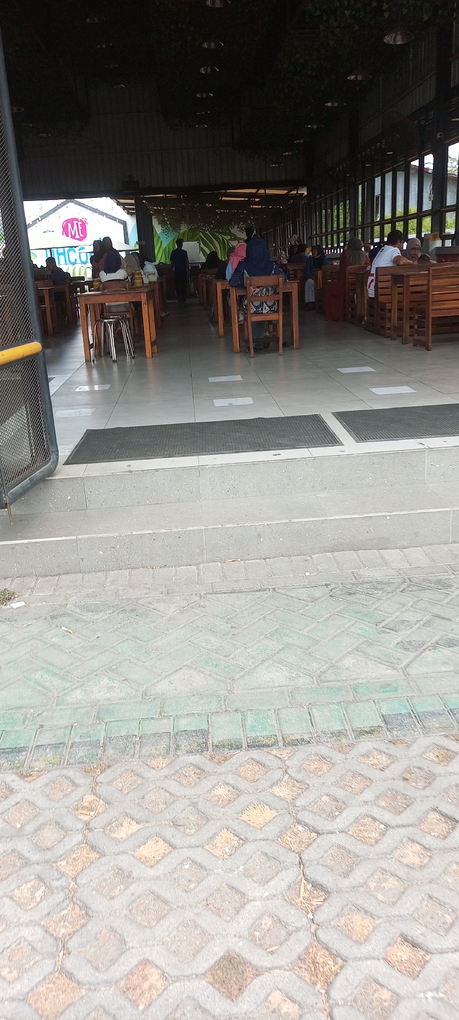 The image depicts an outdoor seating area of a restaurant, likely located in Dapur, Indonesia. Several people are seated at wooden tables and chairs, engaged in various activities such as dining and conversing. The architecture suggests a covered patio or open-air dining space, with a dark ceiling and visible structural beams. A sign with the letters "ME" in pink and white is visible on a building in the background, possibly indicating the name of the establishment. The floor in the foreground is paved with interlocking concrete blocks, some of which are green. There are also steps leading up from the paved area. The overall scene conveys a casual and social atmosphere.