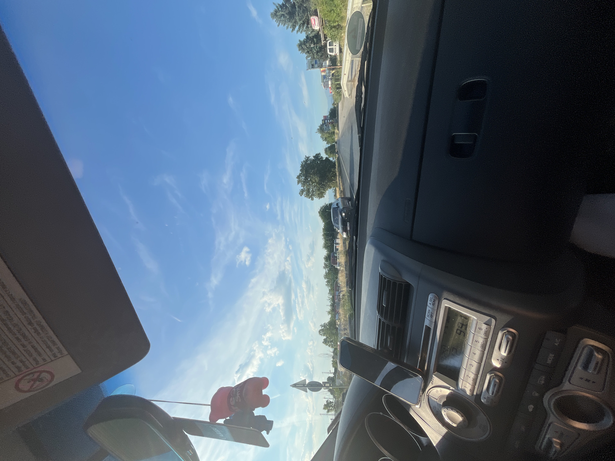 The image shows the interior of a car dashboard looking out at a road. On the dashboard, there is a phone mounted to the windshield, a car radio with visible display numbers, and various buttons. A red plush toy is hanging from the rearview mirror. Outside the car, the road stretches ahead under a bright blue sky with scattered clouds. There are trees and buildings visible along the roadside, with other vehicles on the road. The scene appears to be in Graf Ignatievo, Bulgaria, given the context provided.