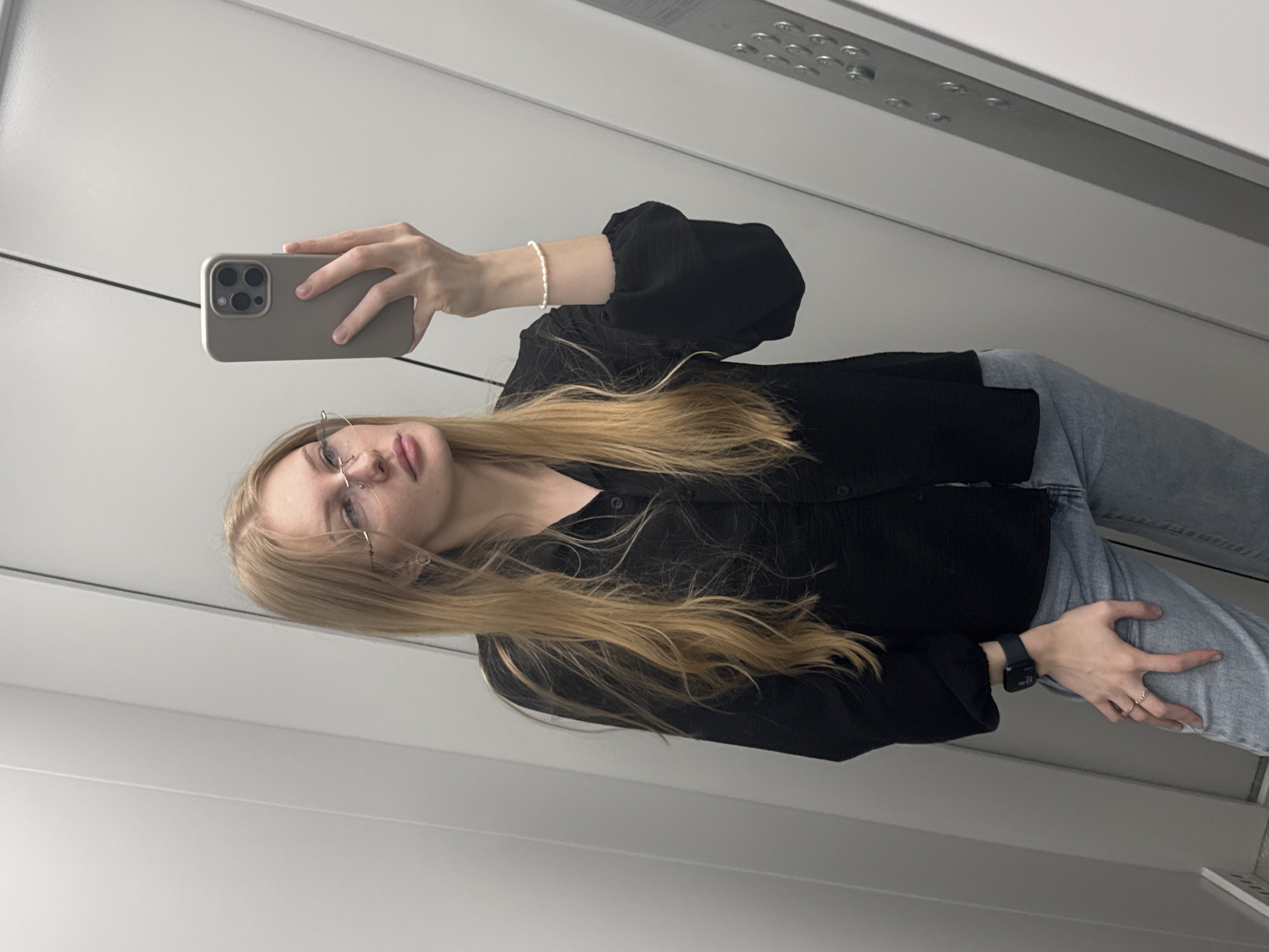 A young woman with long blonde hair and glasses is taking a mirror selfie in what appears to be an elevator. She is wearing a black collared shirt and light blue jeans. She is holding a smartphone in her right hand, and her left arm is adorned with a pearl bracelet. A smartwatch is visible on her left wrist. The background is a plain, light-colored wall, likely part of the elevator interior, with some metallic elements at the top. The lighting suggests it's daytime. Her expression is neutral, and she is looking at the phone's camera. The image is vertically oriented. There is no visible text in the image. The location context is Perm, Russia.