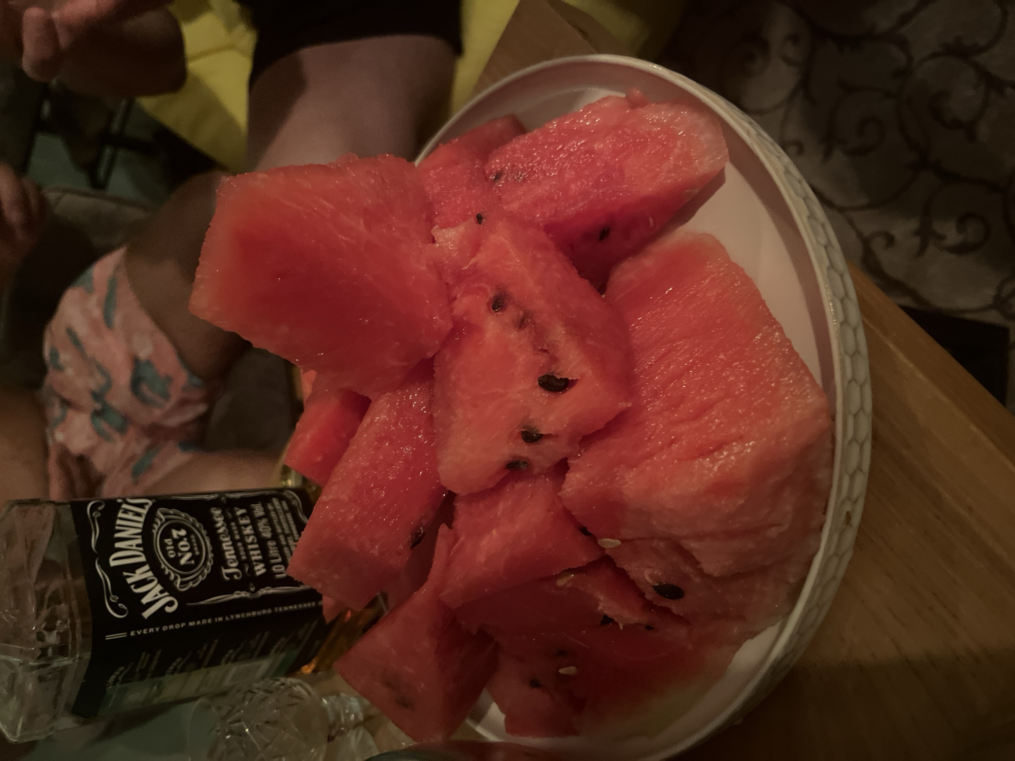 A medium shot depicts a bowl of cut watermelon next to a bottle of Jack Daniel's whiskey. The watermelon pieces are red and contain black seeds. The bottle is partially obscured by the bowl. The background includes an indistinct figure in clothing, a patterned wall, and a wooden surface. There is no mention of the city of Новая Чернушка, Russia, visible in the scene.