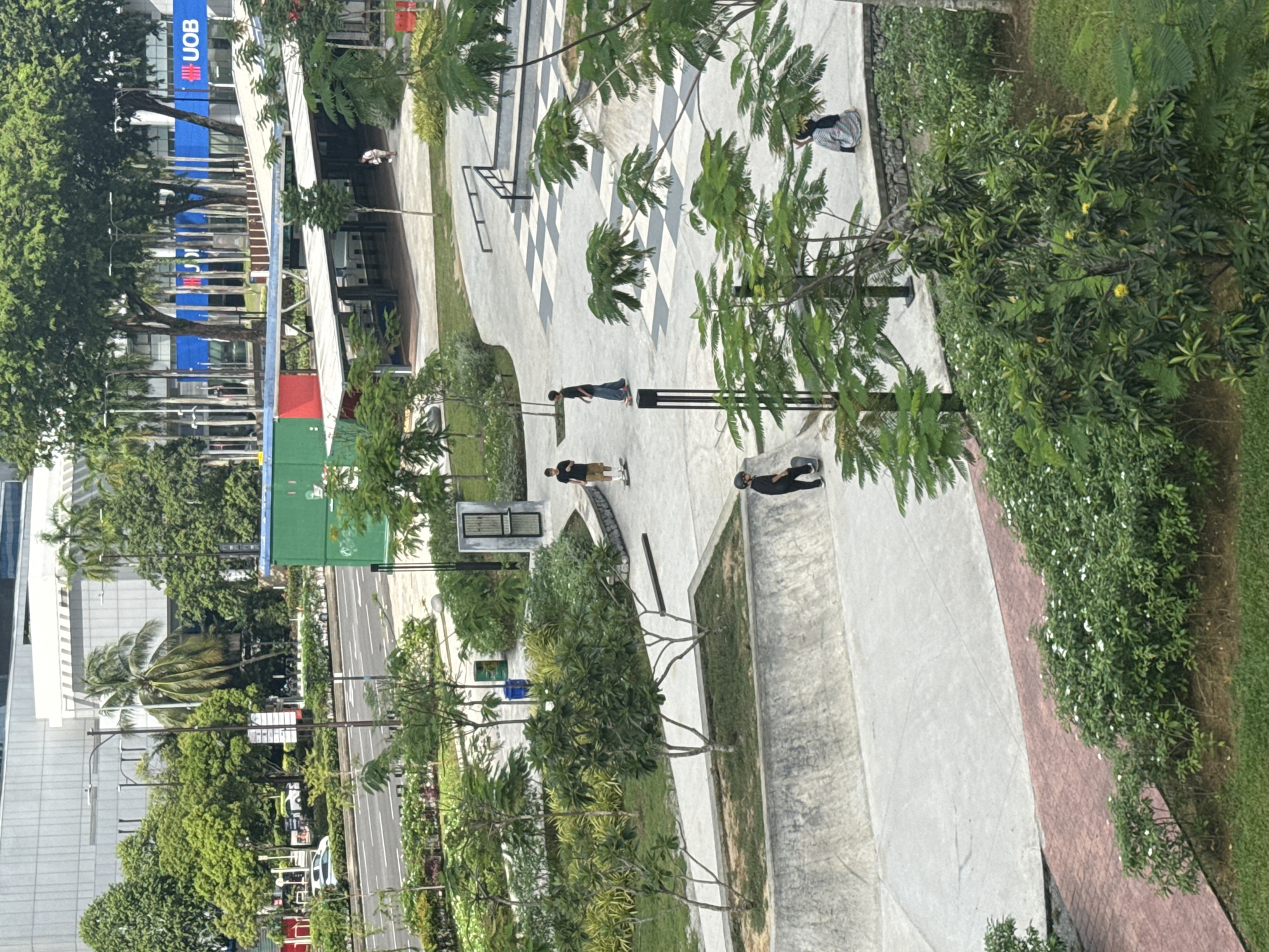 The image depicts an outdoor urban public space, likely a park or a square, in Kuala Lumpur, Malaysia, during bright daytime under clear, sunny skies. The setting features modern concrete pathways, a distinctive checkered grey and white patterned ground area, and abundant greenery including large trees with dense foliage, palm trees, and flowering shrubs.
Several people are present, indicating a casual social gathering or recreational activity. In the midground, three individuals are visible with skateboards, suggesting skateboarding is taking place. One person wears a black t-shirt and dark pants, standing beside a skateboard. Another in a black t-shirt and tan shorts also stands near a skateboard. A third person, dressed in black, is resting on the ground with a skateboard nearby. Further back, a person in a striped headscarf and light-colored top is seen bending or sitting near the checkered pattern.
Visible objects include the skateboards, green public bins, streetlights, and what appear to be street furniture or rails integrated into the concrete design. In the background, a busy street with cars is visible, flanked by modern multi-story buildings. A prominent blue sign on one of these buildings displays the text "UOB," identifying a commercial bank. The bright lighting casts soft shadows, contributing to a relaxed daytime atmosphere.