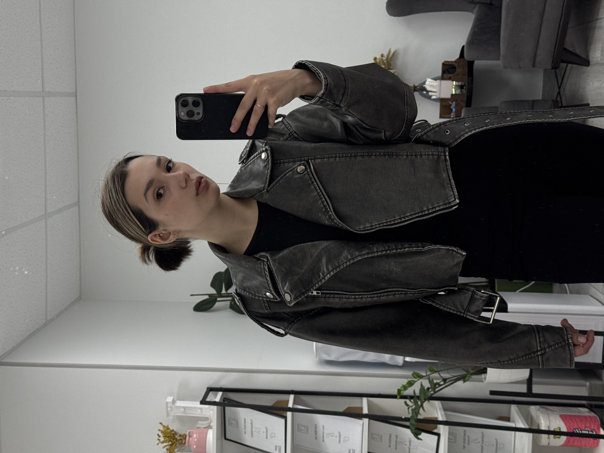 A young woman is depicted in a mirror selfie, facing the camera with a neutral expression. She has light brown hair with blonde highlights, styled in a low bun, and is wearing a dark, distressed grey faux leather moto jacket over a black top. A simple ring adorns her left ring finger as she holds a black smartphone to capture her reflection.
The setting appears to be an indoor space, likely a modern office, studio, or commercial establishment, indicated by the white walls, a white ceiling with acoustic tiles, and artificial lighting. In the background, a grey upholstered armchair and possibly another seating unit are visible, along with a reflective surface or small table.
In the lower portion of the image, a white shelving unit with black framing holds several framed documents or certificates. Partially visible text on these frames includes words such as "SCHEMES," "DESIGN," "YOUR D...", "COLLECTION," and the year "2023," suggesting a professional context perhaps related to design or achievements. A small plant with broad green leaves sits on the shelf, alongside a pink ceramic vase containing dried yellow flowers. A roll of paper towels with pink packaging is also partially visible at the bottom right.
The overall ambiance is clean and modern, with functional decor. The time of day is indeterminate due to the indoor setting and artificial lighting.