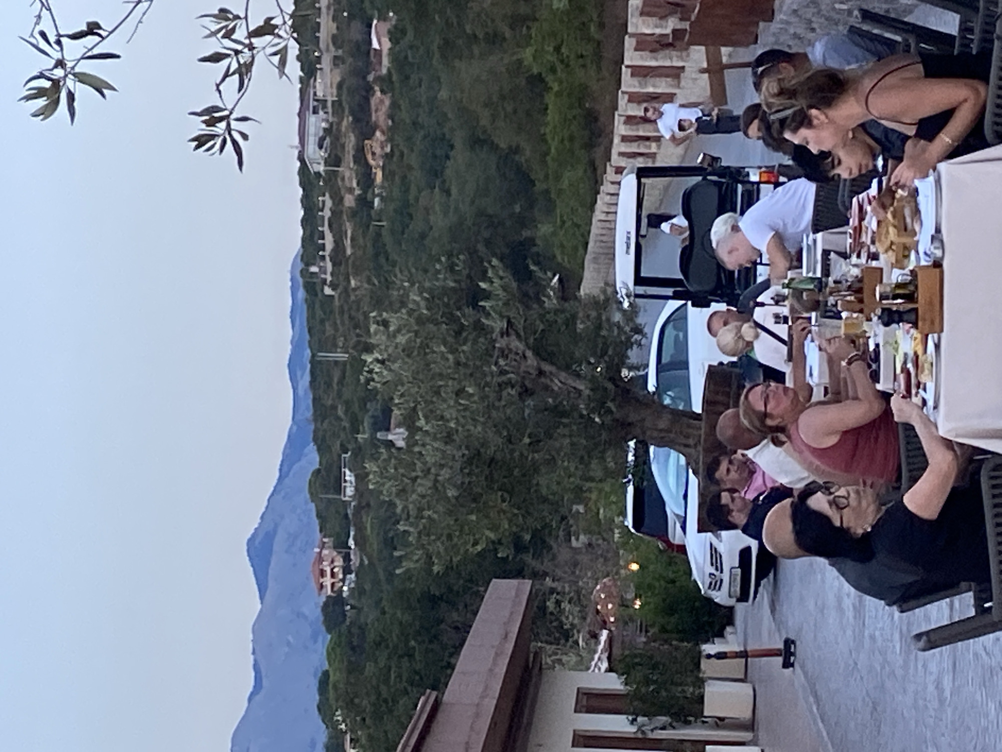 In Kovačko Polje, Montenegro, a group of people are dining outdoors at tables set with food and drinks. The scene appears to be a restaurant or outdoor dining area, with a golf cart visible in the background. Several individuals are seated at tables, engaged in conversation and eating. The environment is scenic, with hills and mountains in the distance, and trees scattered throughout the landscape. The lighting suggests it is either early morning or late evening.