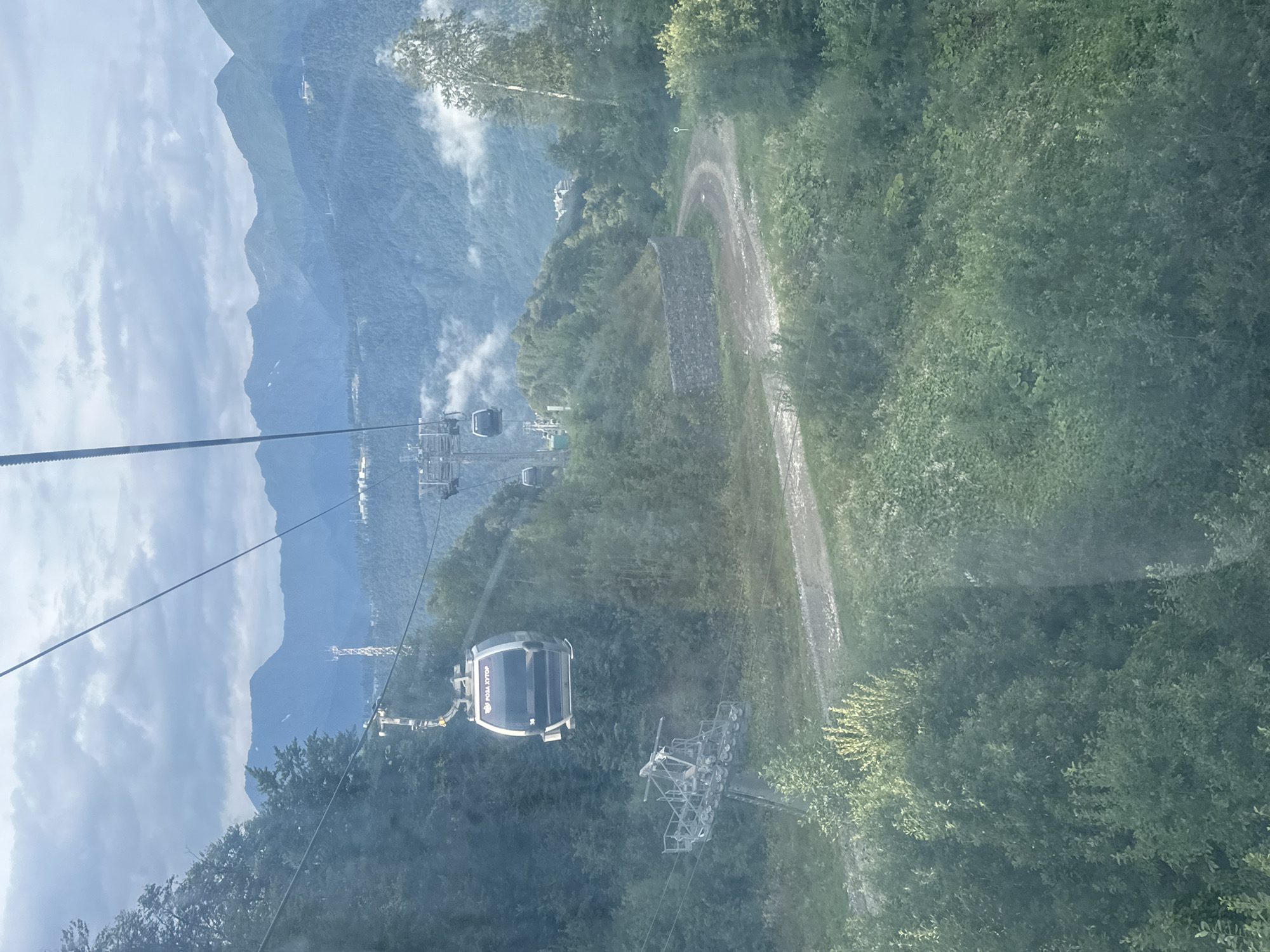 The image captures a view of the Rosa Khutor ski resort in Estosadok, Russia, from a cable car. Multiple cable car cabins are visible suspended from wires, with one prominent cabin in the foreground displaying the text "РОЗА ХУТОР". The surrounding environment is mountainous and densely forested with lush green trees. A dirt road winds through the landscape, bordered by a stone retaining wall. In the background, layers of mountains extend into the distance under a cloudy sky. The scene suggests a summer or late spring day, as the foliage is in full bloom.