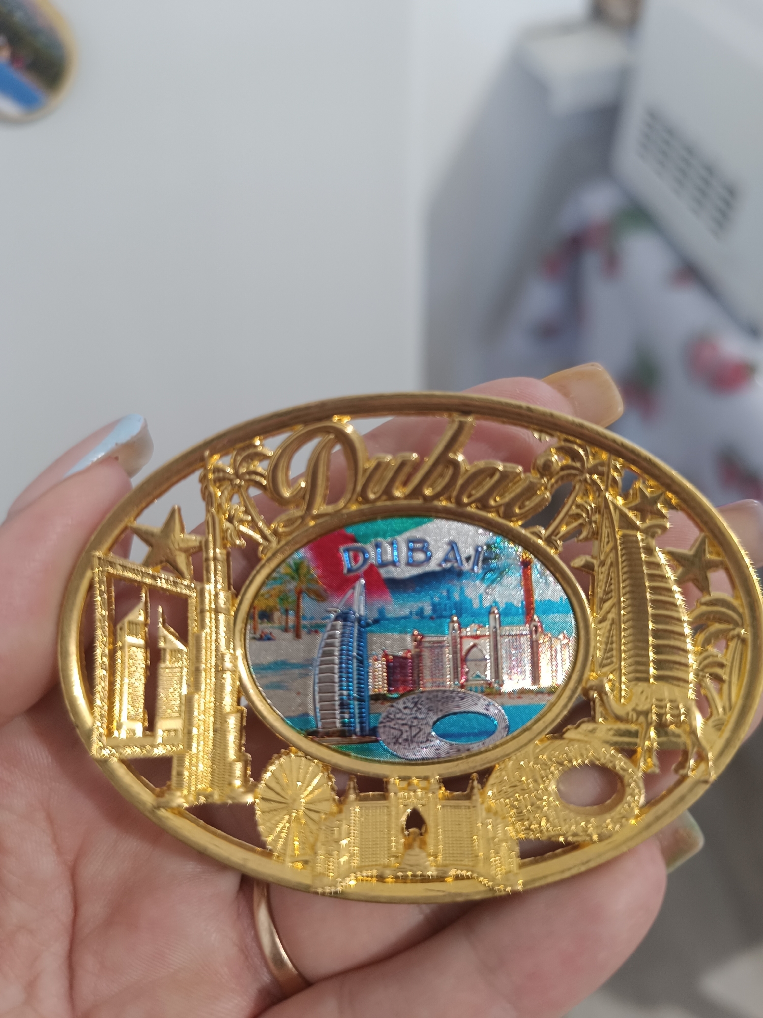 A person is holding a gold-colored oval-shaped souvenir magnet that depicts a scene of Dubai. The magnet features a cutout of the word "Dubai" at the top, with an oval-shaped inset in the center showing various landmarks of the city. These landmarks include skyscrapers, palm trees, and a sail-shaped building. The word "DUBAI" is also visible in blue text within the inset. The souvenir is intricately designed with decorative patterns and cutouts around the edges. The person's hand, with painted fingernails and a ring on one finger, is visible supporting the magnet. The background is blurred and appears to be an indoor environment with a white wall. There is no visual information within the image that relates to the city of Taraz, Kazakhstan.
