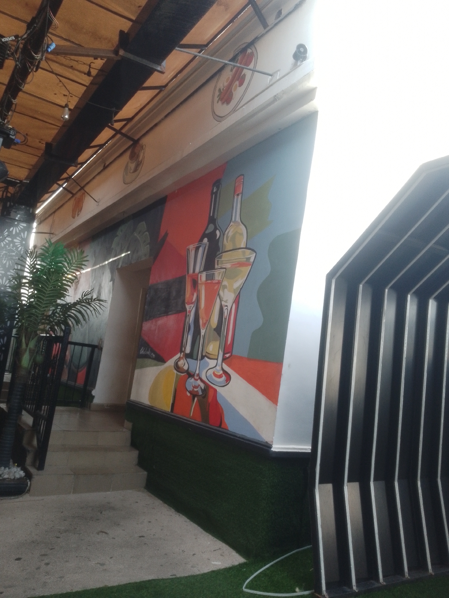 The image depicts an outdoor area, possibly a patio or walkway, with a distinctly artistic mural on a wall. The mural features stylized representations of wine bottles and glasses, rendered in vibrant colors against a muted background. To the left, a potted palm tree adds a touch of greenery. A set of stairs leads upwards, with a concrete pathway at the bottom. The area is bordered by artificial turf. In the background, a corrugated metal structure with a ribbed design is visible. The overhead structure suggests a covered walkway or canopy, supported by wooden beams and metal framework, with visible electrical wiring. The bright sunlight indicates it is daytime. The scene appears to be part of an establishment with a focus on a relaxed, possibly dining or social, outdoor experience. The location is identified as Howayi, Nigeria.