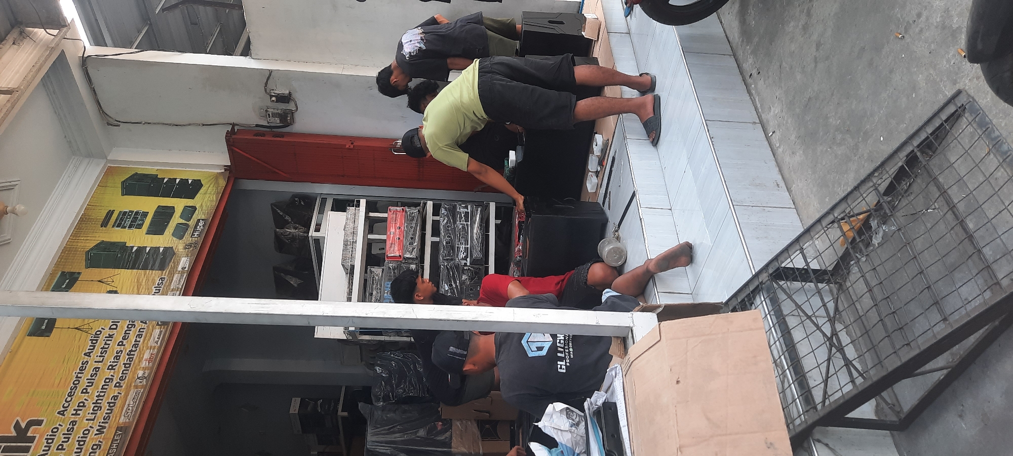 In Selopuro, Indonesia, outside a shop with a banner advertising audio and accessories, several individuals are engaged in an activity involving large black speaker cabinets. One man in a green t-shirt and black shorts is positioned behind a speaker cabinet, appearing to adjust it. Another man in a black t-shirt with a graphic print and dark shorts stands nearby, facing away from the camera, and a third man in a red shirt and dark shorts is seated on the ground. A fourth individual, wearing a black t-shirt with "GL LIGHTING PROFESIONAL" and a WA number printed on it, is also seated. The environment consists of tiled steps leading up to the shop entrance, with various speaker cabinets and electronic equipment stacked on shelves inside the shop. A metal ramp with a wire mesh surface is present in the foreground, and discarded cigarette butts are visible on the ground outside the shop. The overall scene suggests a commercial setting where audio equipment is being handled or arranged.