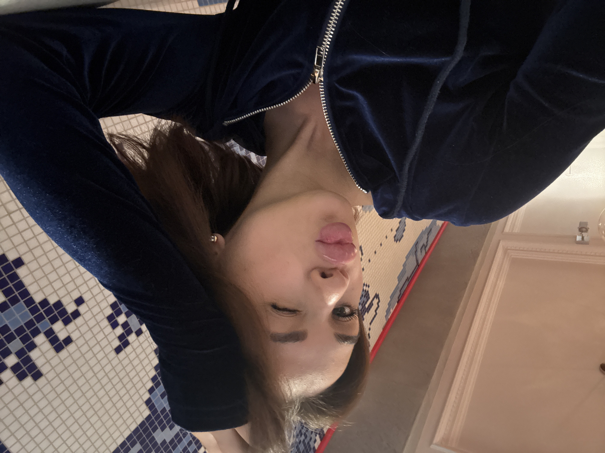 This media file features a young woman posing playfully in an indoor setting in Perm, Russia. She is shown from the chest up, with her head slightly tilted and her right eye winking while her tongue is playfully sticking out. Her lips appear glossy, and her makeup includes defined eyelashes and eyebrows. She has long, dark brown hair and wears a small, silver stud earring in her visible ear.
She is dressed in a dark blue velour or velvet tracksuit jacket, which has a prominent silver zipper that is partially undone, revealing a glimpse of her neck. The background to her left consists of a floor or wall covered in small mosaic tiles, creating an abstract pattern with white and various shades of blue squares. A thin red line separates this tiled surface from a plain, light-colored wall or ceiling on her right, which features decorative molding near the top. The lighting suggests an artificial, indoor environment. There is no visible text in the image.
