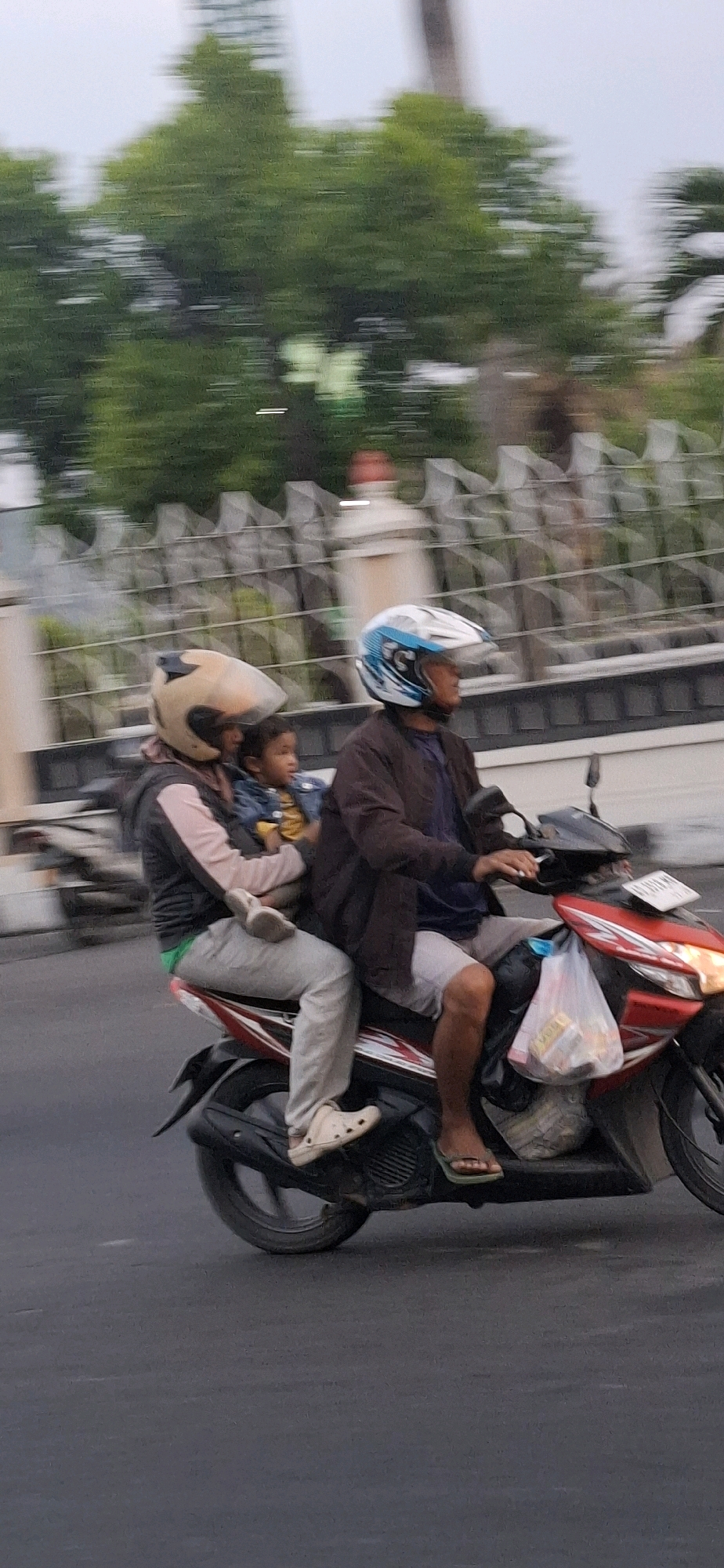 Three people are riding on a red and black motorcycle on a paved road in Desa Donohudan, Indonesia. The person in the front is wearing a brown jacket and a blue and white helmet. They are driving the motorcycle. The person in the middle is wearing a beige helmet and a pink and black jacket, and they are holding a child. The child is wearing a blue jacket over a yellow shirt. The person in the middle is wearing gray pants and white clogs. The person in the front is wearing gray shorts and flip-flops. There is a plastic bag with items inside hanging from the front of the motorcycle. Trees and a fence are visible in the background. The image is blurry due to motion.