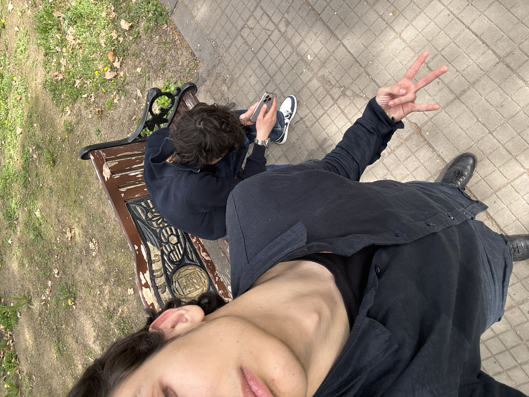 Two young men are photographed from above in Yerevan, Armenia. One sits on a weathered park bench with peeling paint, looking at a smartphone. He wears a dark hooded sweater, jeans, and white sneakers. The other man stands with his arm raised, making a "W" or "peace" sign with his fingers. He wears a dark shirt and dark shoes. The ground is paved with square tiles, and there is grass and foliage near the bench.