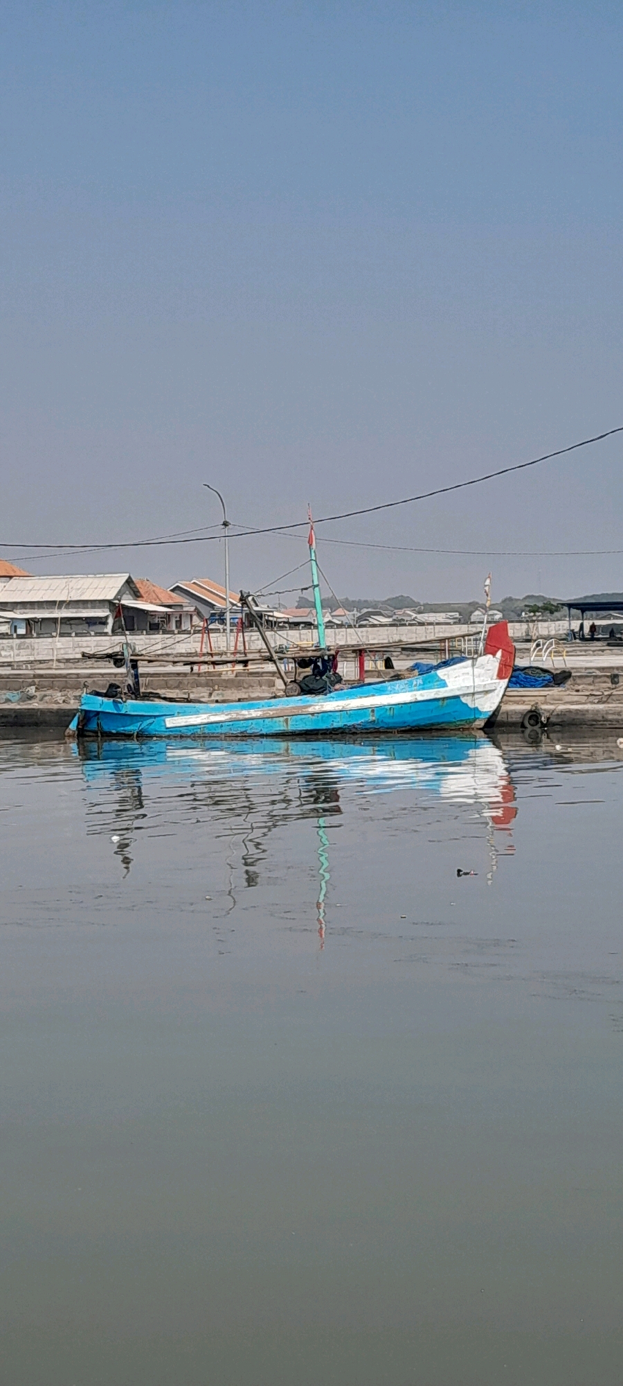 A weathered, blue and white fishing boat rests at anchor in the murky waters of the Pasuruan River, Indonesia. The boat, with its distinctive red and white flag on a tall mast, is tethered to a concrete embankment lined with buildings. The buildings appear to be a mix of residential and commercial structures, with corrugated metal roofs and pale walls. A few people are visible on the embankment, some tending to the boat, while others are simply present. The water's surface is still, reflecting the boat and the surrounding structures, creating a distorted yet recognizable mirror image. The sky above is a clear, pale blue, indicating a bright, possibly hot, day. The scene depicts a typical day in a riverside community, where boats and water are integral to daily life.