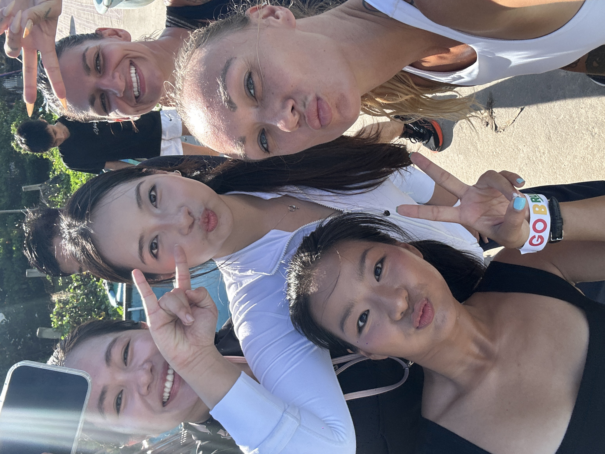 Five individuals, four women and one man, are captured in a close-up, outdoor selfie on a sunny day. The foreground is dominated by the faces of four women, positioned in a tightly grouped selfie. One woman, on the left, holds a smartphone and smiles broadly while making a peace sign with her left hand. Next to her, another woman is puckering her lips and her left hand is raised with fingers extended. To the right, a third woman with blonde hair is also puckering her lips, her eyes looking towards the camera. In the bottom right quadrant, a fourth woman with dark hair is leaning into the frame, her lips pursed. A fifth individual, a man wearing a black t-shirt and white shorts, is visible in the background, looking down.
The background suggests an outdoor environment with some greenery and a fence or barrier. The ground appears to be a paved surface. The overall impression is a casual, social gathering. It is not possible to definitively determine the location as Mũi Né, Vietnam, based solely on the visual information provided.