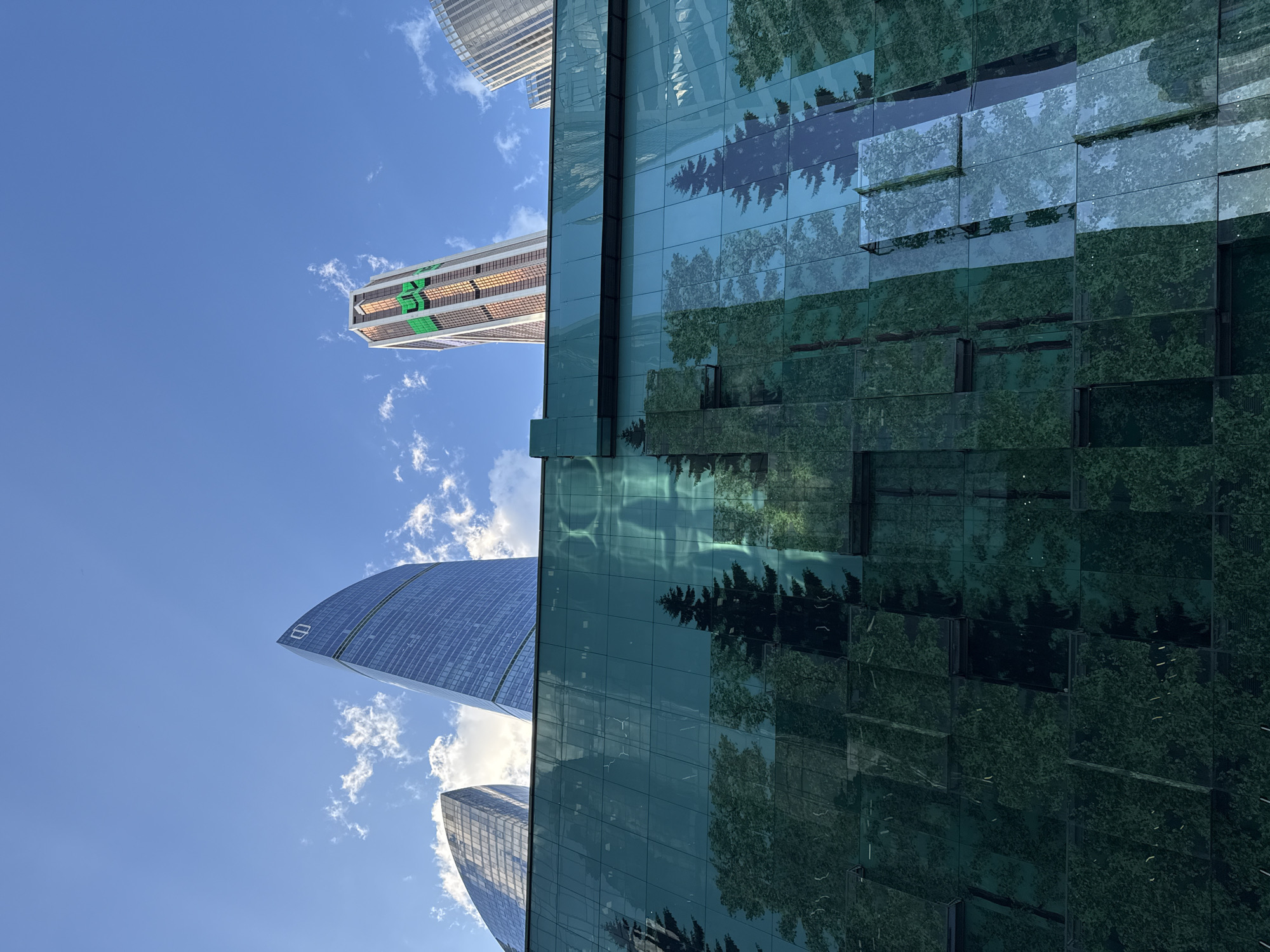 This vertically oriented image captures a low-angle perspective of several modern skyscrapers under a bright blue sky with scattered white clouds, emphasizing their height and glass facades.
In the foreground, dominating the right half of the frame, is a large contemporary building clad in a dark green, reflective glass facade. This surface is segmented into numerous rectangular panels, creating a mosaic effect. These panels prominently reflect the dark silhouettes of evergreen trees and sections of the blue sky, with reflections varying in brightness and clarity.
To the left, a tall, sleek skyscraper with a blue-tinted glass exterior curves upwards. Near its top, a white, stylized logo resembling an "8" or an abstract "B" is visible. Further back, another skyscraper with a lighter-colored, possibly white or light grey, facade features horizontal banding. On one side of this building, large, rectangular green patterns are visible, which appear to form a stylized logo or architectural design, possibly resembling "S3" or "E3". The top edge of another light-colored building is partially seen at the very top of the image.
The scene depicts a clear, sunny daytime environment. No specific human activity is occurring. The combination of reflective surfaces, imposing height, and distinct architectural features underscores a modern urban landscape.