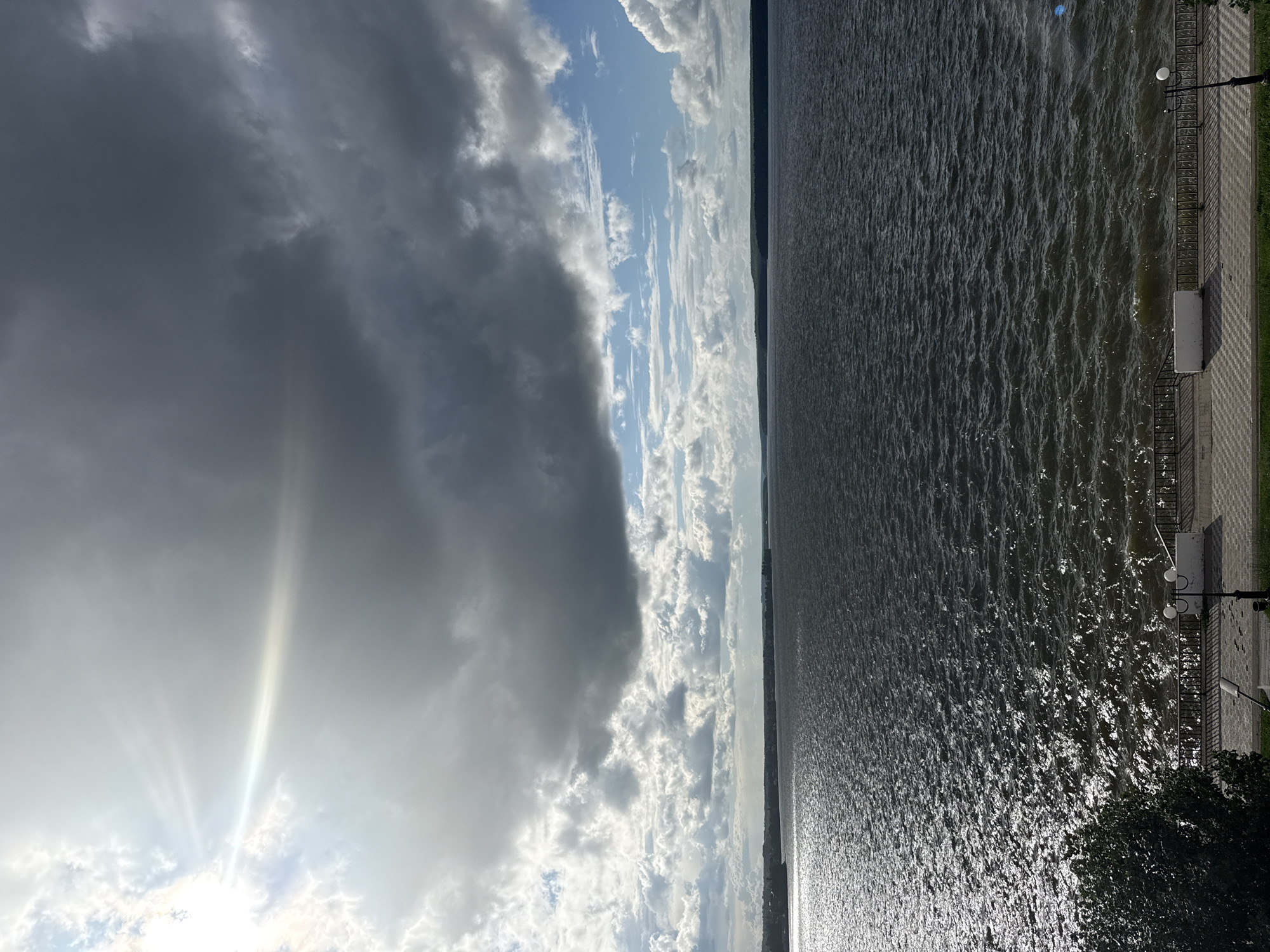 This is a vertical, wide-angle photograph taken from a slightly elevated position, looking out over a body of water. The image is divided into two distinct halves by the horizon. The left side is dominated by a dramatic sky filled with a mix of dark, heavy clouds and brighter, fluffy white clouds. Sunlight pierces through the darker clouds in a strong, visible ray effect, suggesting either dawn or dusk, or a break in stormy weather. The right side of the image shows a vast expanse of dark, rippling water, likely a lake or river, reflecting the light from the sky in shimmering patterns.
Along the edge of the water, on the far right, is a paved walkway or embankment with a decorative metal railing. A lamppost with two globes stands near the railing, and a patch of green grass is visible behind it. A small bench is also present along the walkway. In the foreground on the right, a dark green tree branch enters the frame, partially obscuring the walkway and water. In the distance, along the horizon line between the sky and water, a faint outline of land or buildings is visible, suggesting an urban or semi-urban setting.
The overall mood of the image is one of dynamic natural beauty, with the contrast between the stormy clouds and the bright sunlight on the water creating a sense of awe. The time of day appears to be either early morning or late afternoon, given the quality of light. The location cues, such as the embankment and lamppost, are typical of a waterfront promenade. There is no visible text in the image.